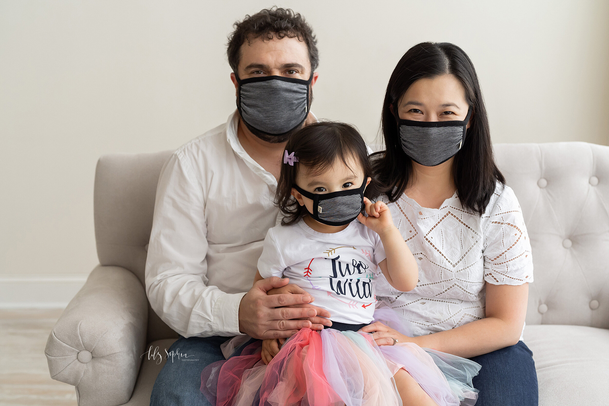  Family photo of a mom, dad, and their two-year old daughter as they sit on a sofa in a natural light studio sporting their masks during the corona virus pandemic in the Poncey-Highlands area of Atlanta. 