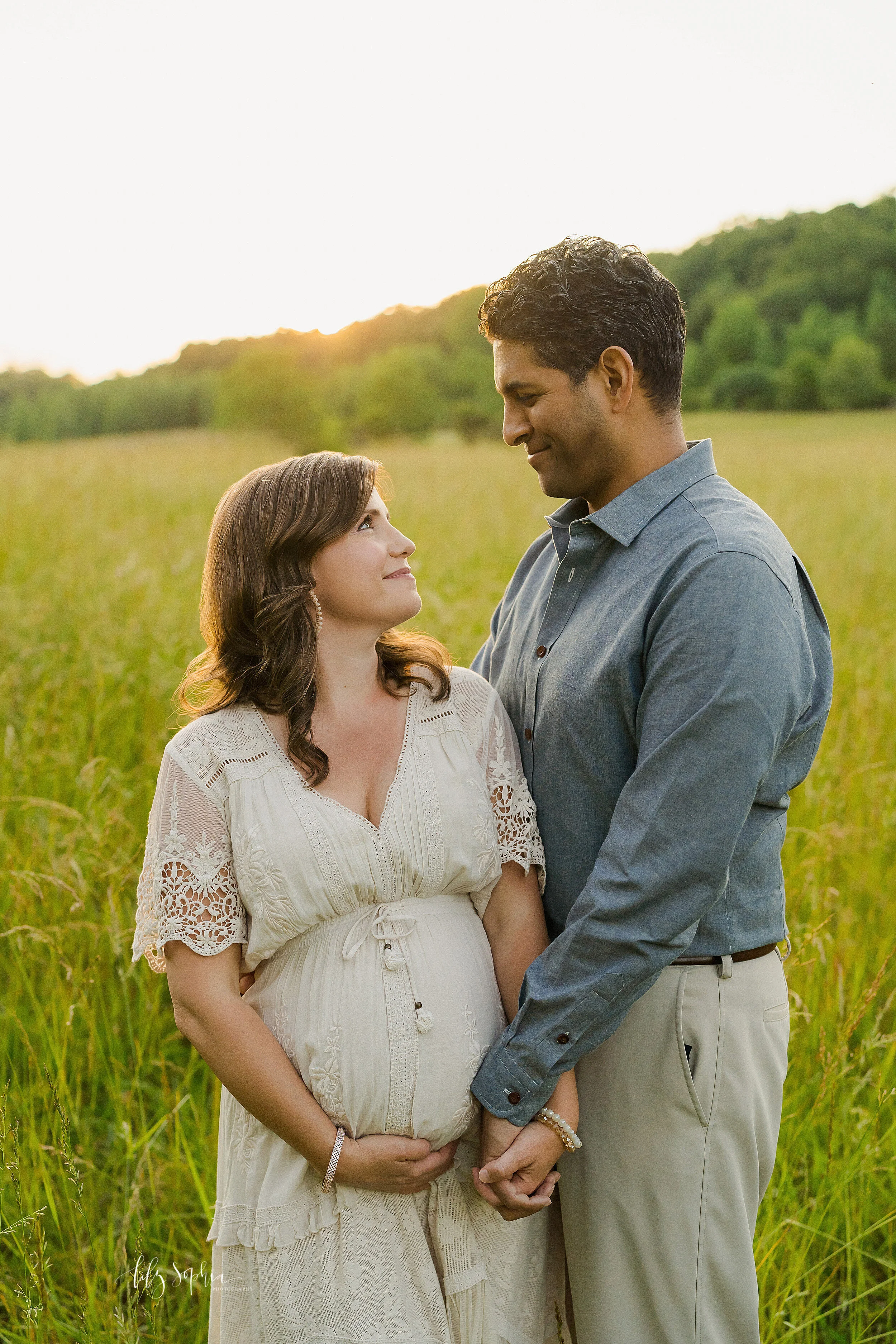  Maternity photo of a husband and wife standing in a field looking lovingly at one another while the sun sets in the background near Atlanta, Georgia. 