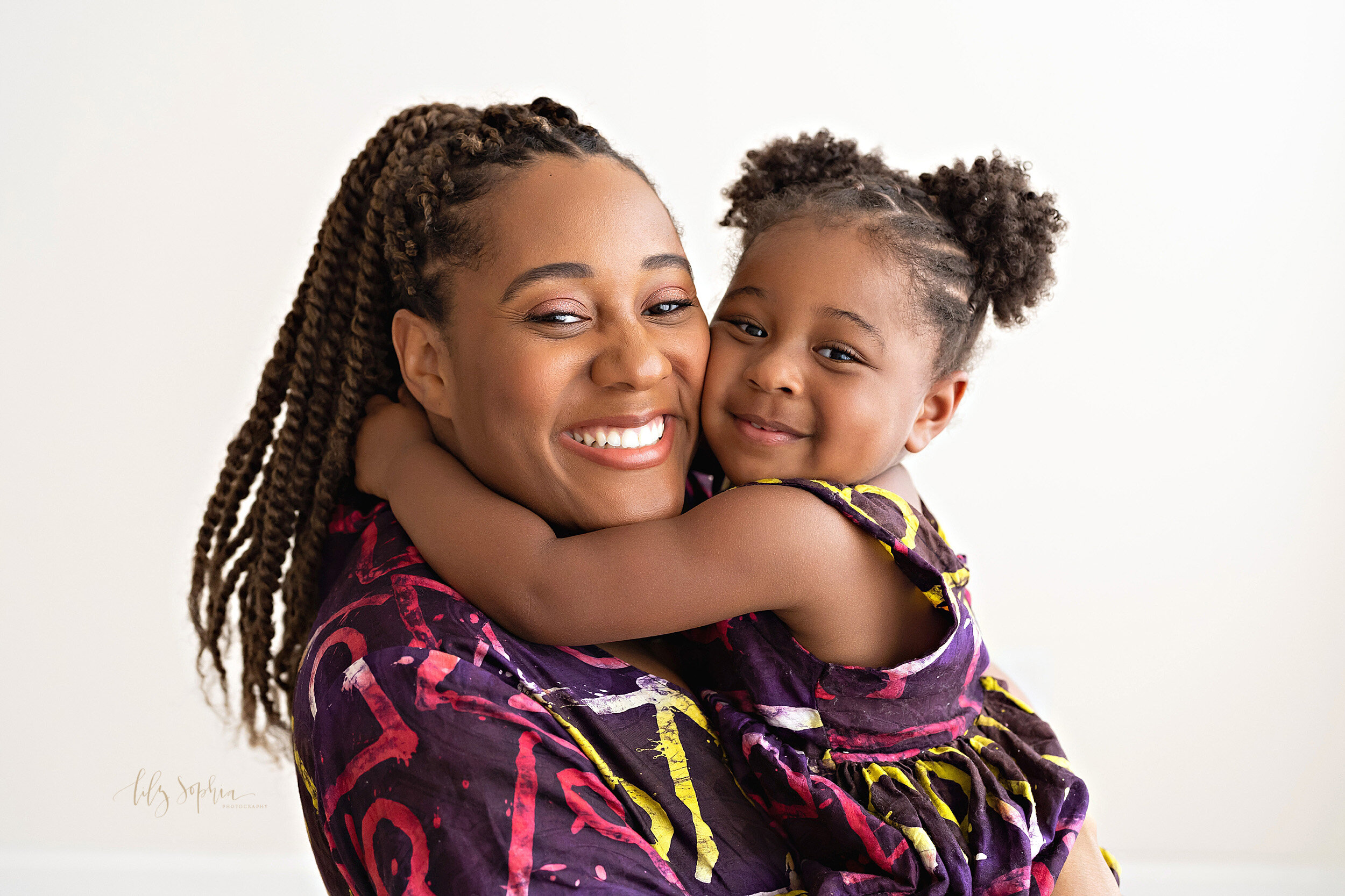  Family picture of an African-American mom standing as she holds her toddler daughter and her daughter hugs her around her neck taken in the Ponce City Market studio of Lily Sophia Photography in natural light. 