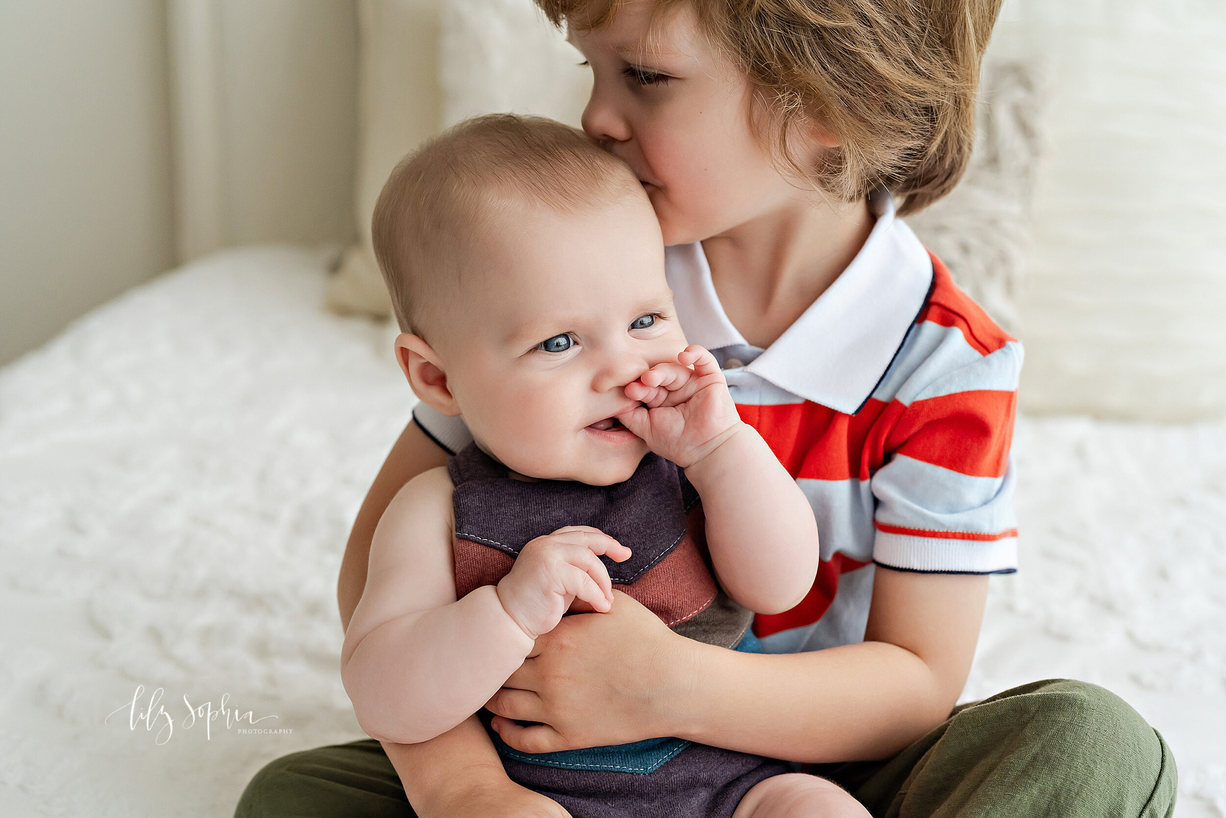  Sibling photo of a toddler son holding his six month old sister on his lap on top of a bed with the older brother kissing his sister on the head taken in a studio near the Morningside area of Atlanta in natural light. 
