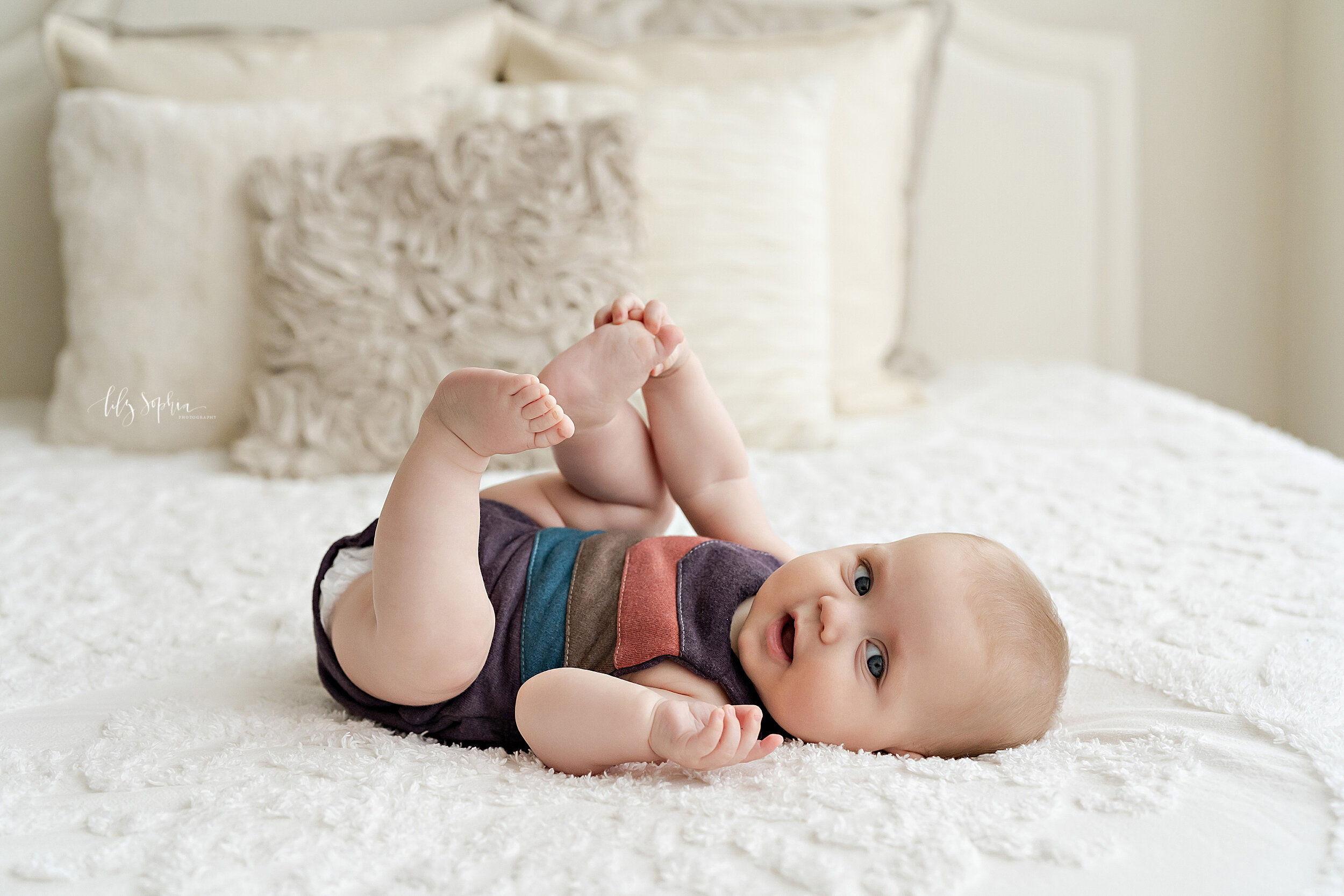  Baby picture of a six month old little girl as she lies on her back on a bed and plays with her toes taken in natural light near the Roswell area of Atlanta, Georgia. 