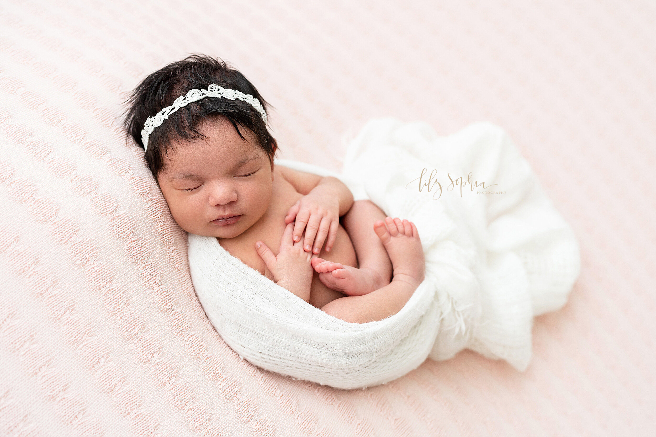  Newborn photo of a peacefully sleeping infant girl wearing a crocheted white headband in her dark hair as she lies on her back on a pink blanket with her hands on her chest and her body cradled in a stretchy white blanket. 