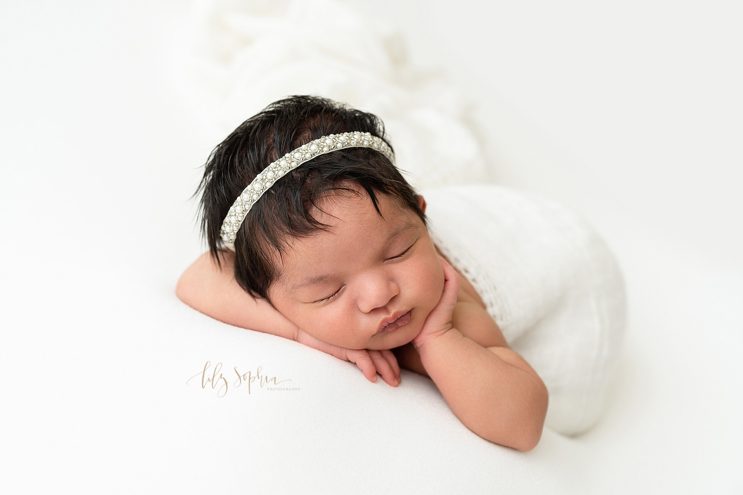  Newborn photo session with a sleeping baby girl lying on her stomach  with one hand under her head and wearing a pearl headband while her other hand rests on her cheek taken in natural light in a studio near the Old Fourth Ward area of Atlanta, Geor