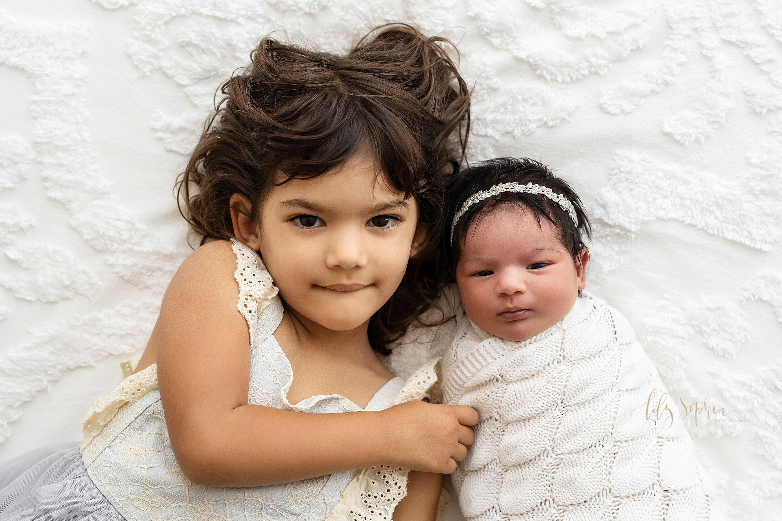  Sibling photo shoot with a toddler girl and her newborn sister as they lie on their backs next to one another on a bed in a studio near the Ansley Park area of Atlanta in natural light. 