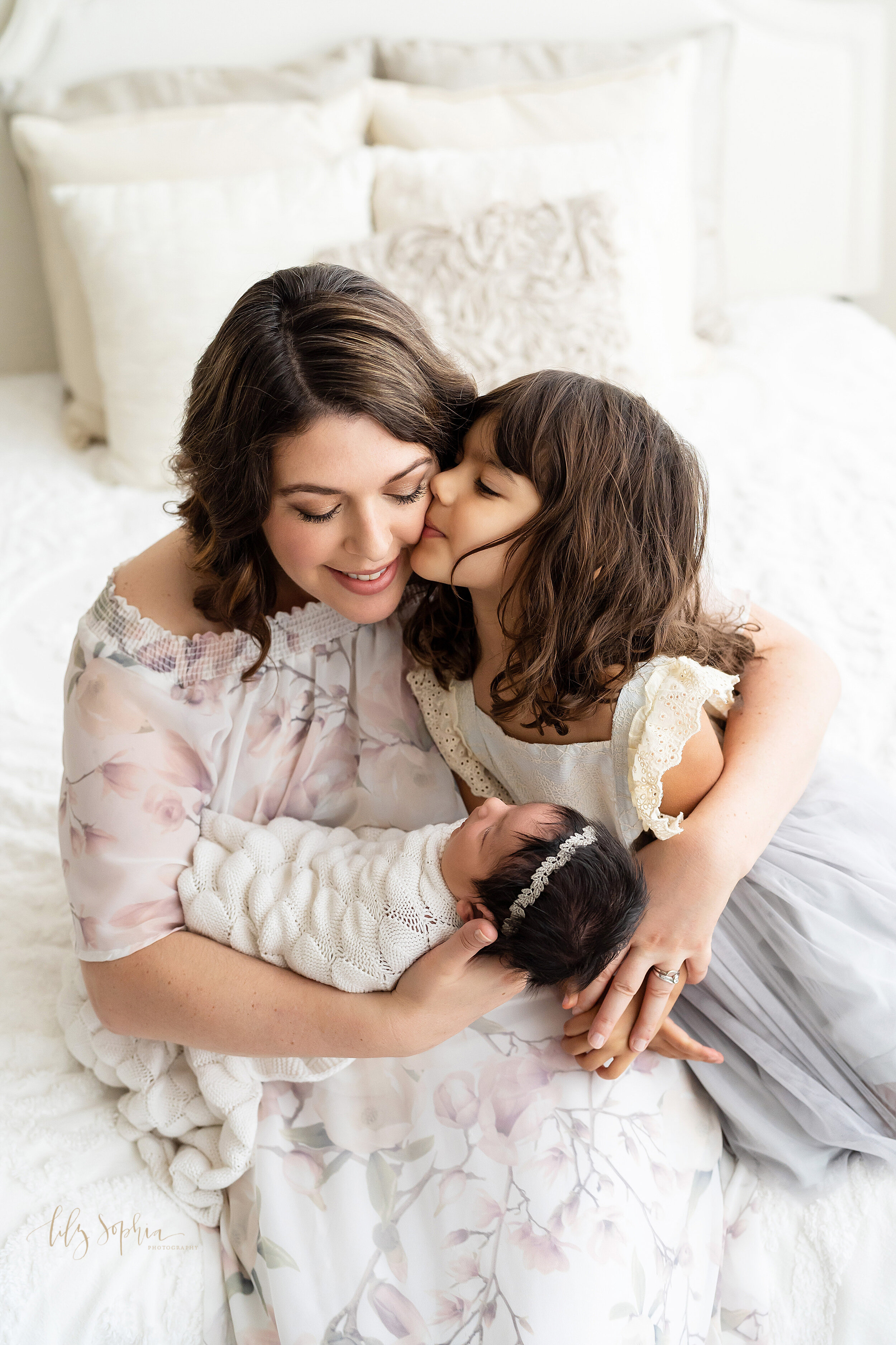  Family newborn picture of a mom and her girls as she sits on a bed in front of a natural light window and holds her newborn daughter in one hand as her toddler daughter kisses her cheek taken near the Candler Park area of Atlanta in a studio. 