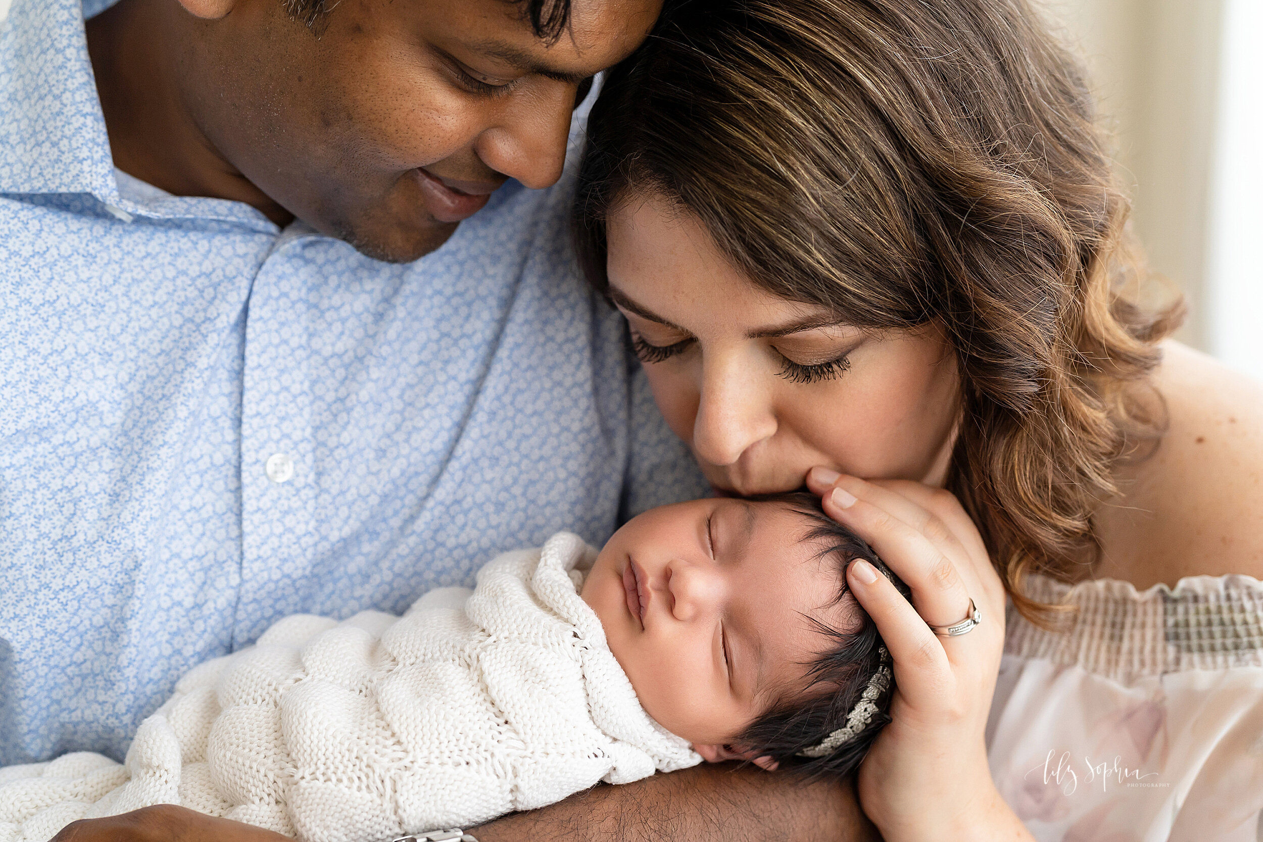  Family photo of a newborn being held in her father’s arms as mom kisses her sleeping daughter’s head taken in a studio near the Midtown area of Atlanta, Georgia in natural light. 