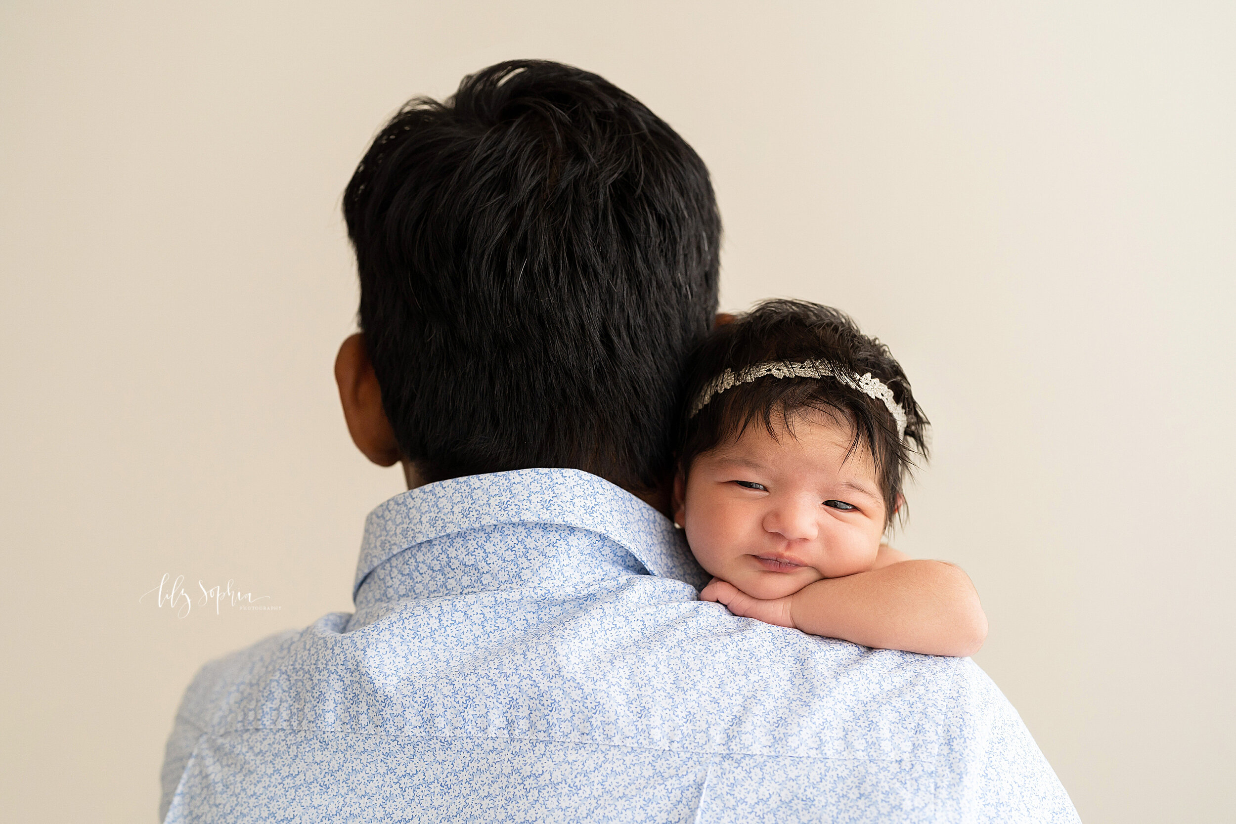  Newborn photo of an infant girl as she peers over her father’s shoulder taken near the Alpharetta area of Atlanta in a natural light studio. 