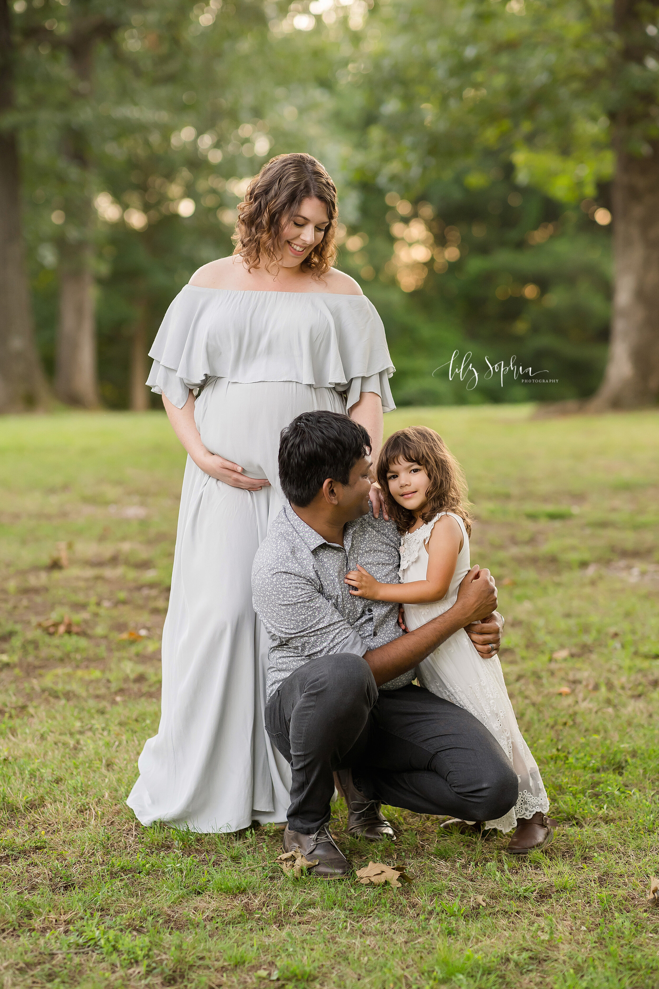 Family maternity photo of a mom dressed in an off-the shoulder full-length gown with her husband squatting in front of her holding their young daughter beside him taken in a park near Atlanta at sunset. 