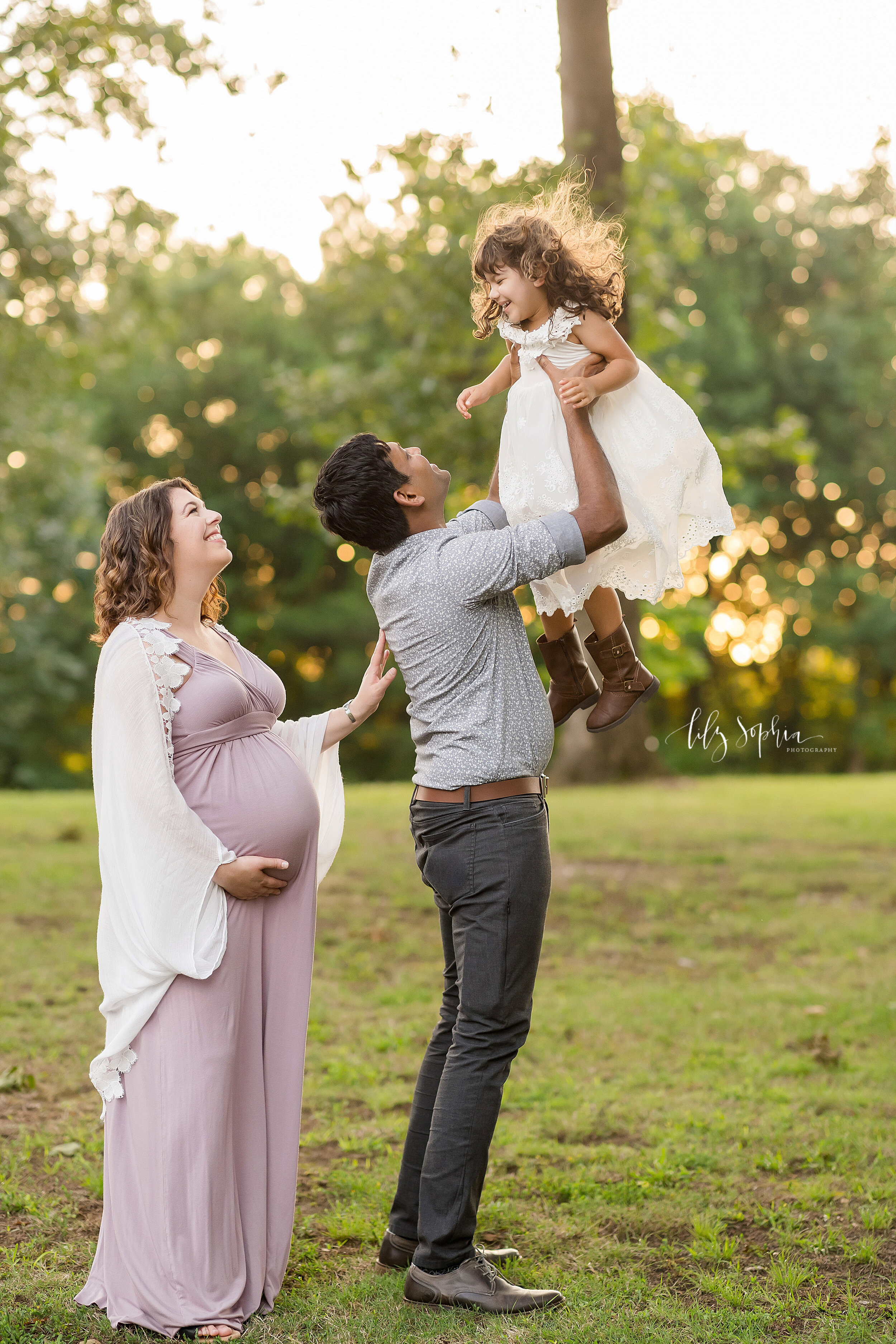  Maternity picture of a family as mom stands behind dad and dad tosses their young daughter into the air taken at sunset in a park near Atlanta, Georgia. 