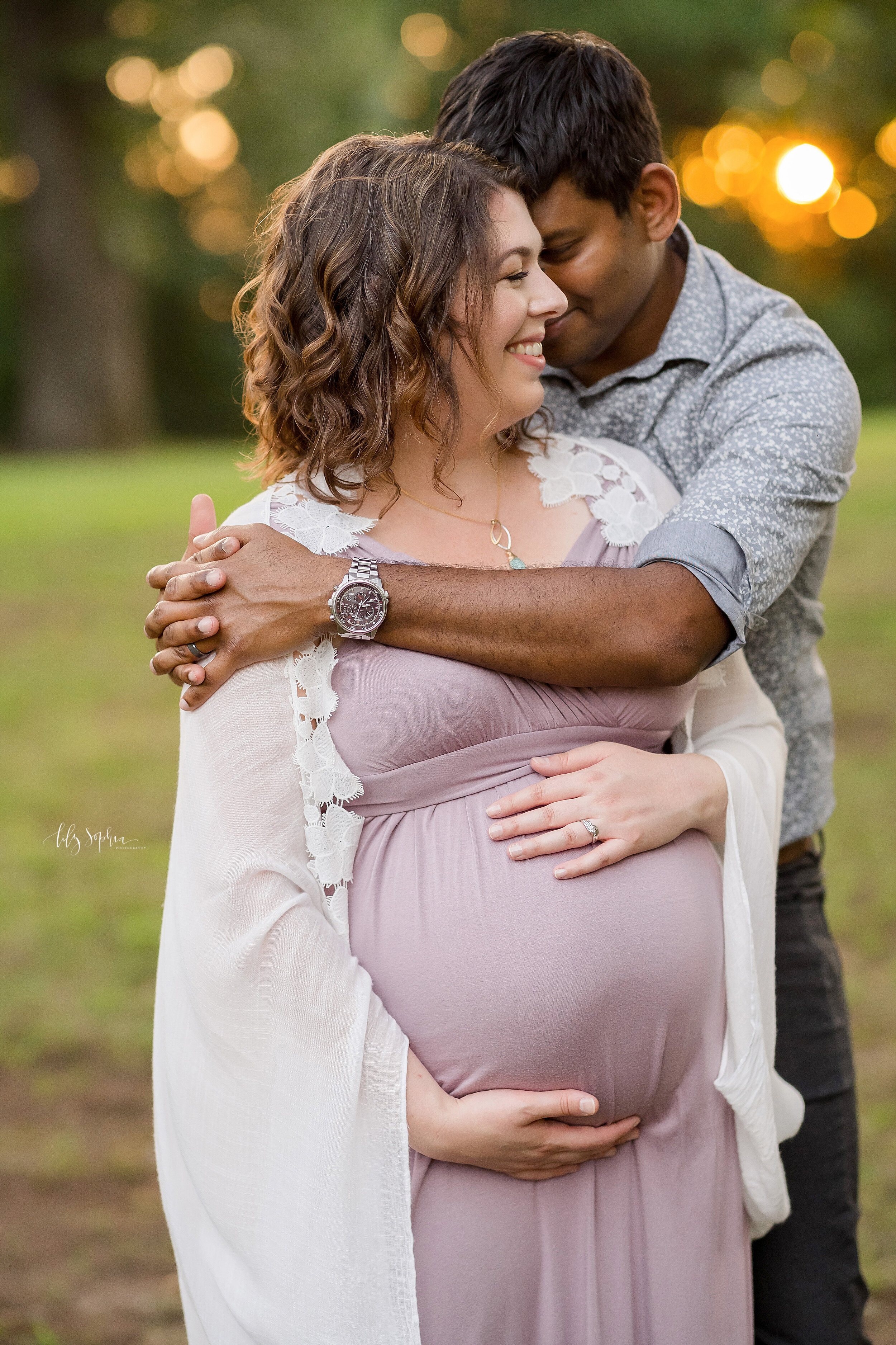  Maternity photo session in a park near Atlanta at sunset with the husband wrapping his arms around his pregnant wife’s shoulders as she frames her belly with her hands. 