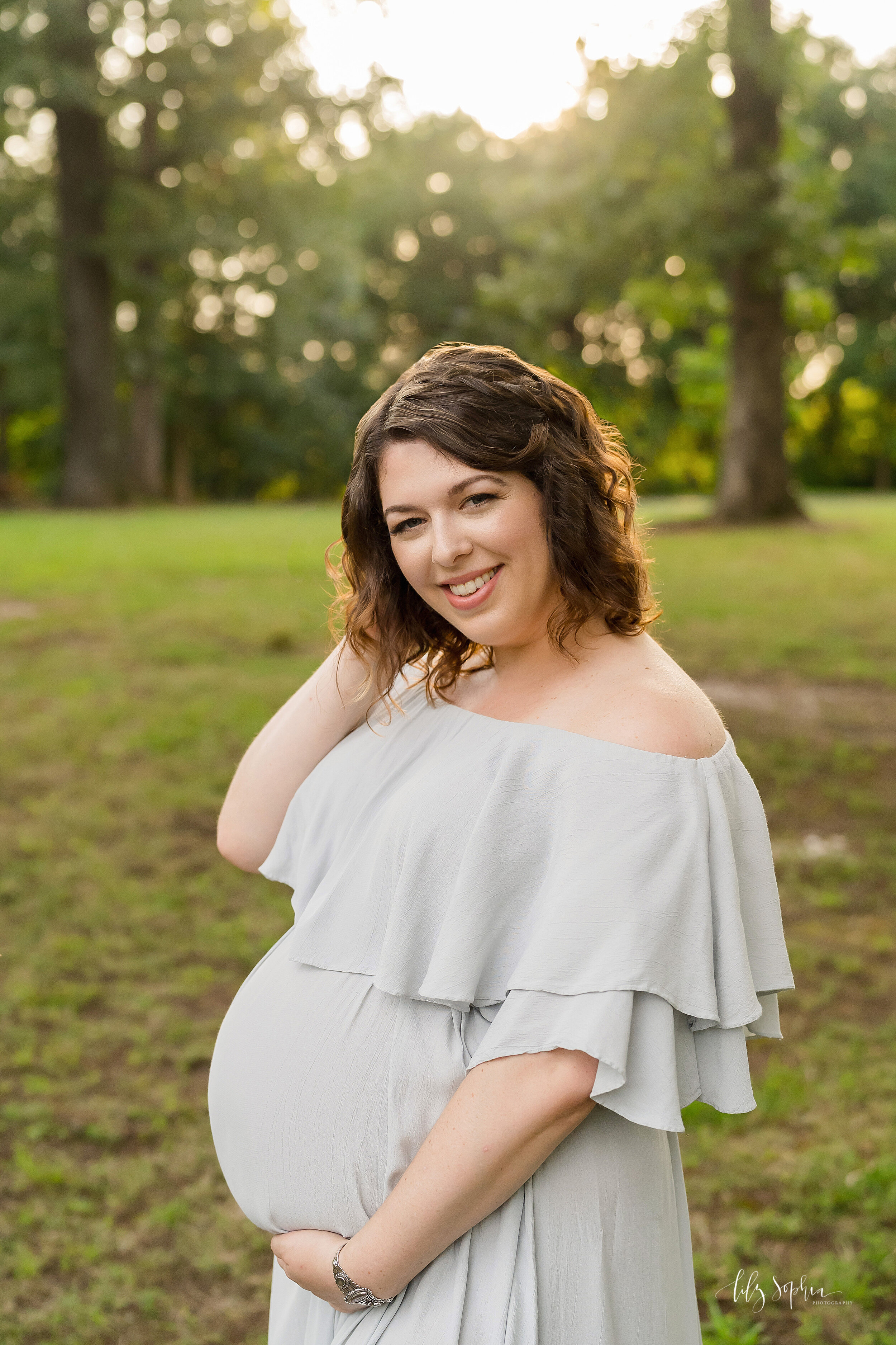  Maternity photo shoot of a pregnant mother dressed in a flowing full-length gown as she stands in a park near Atlanta, Georgia with the sun setting in the background. 