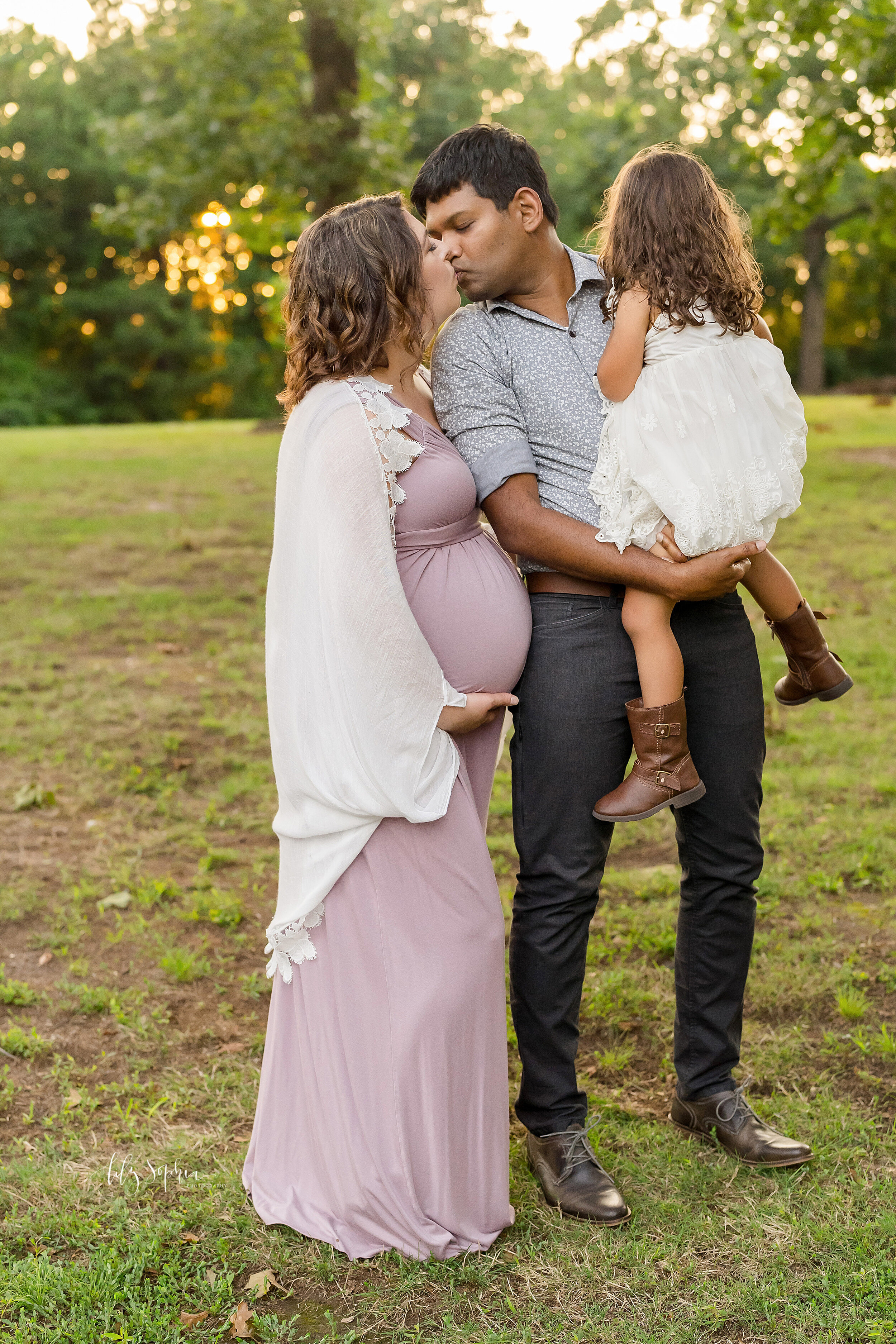  Maternity family picture with mom dressed in a jersey knit gown and a white knit shawl standing next to her husband who is holding their toddler daughter in his arms as he turns over his right shoulder to kiss his pregnant wife as the sun sets in a 