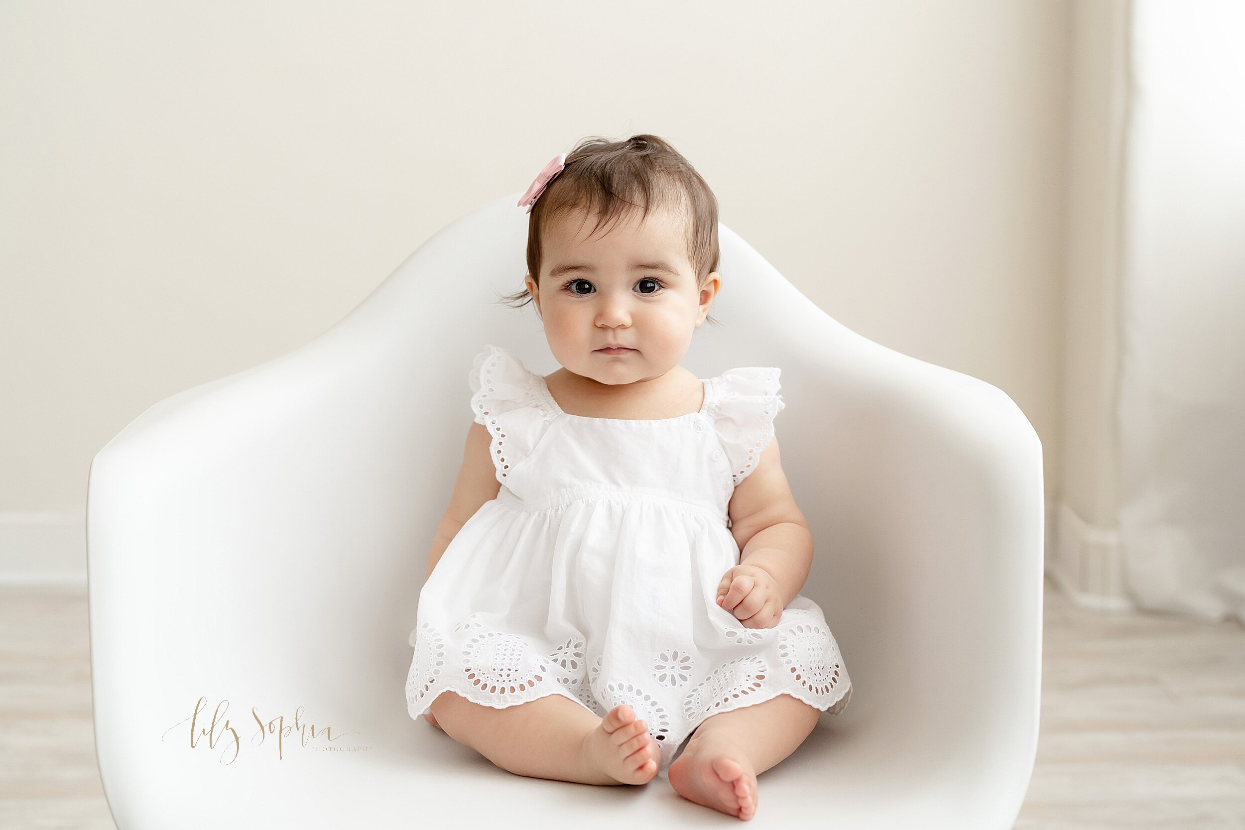  Baby photo of a one year old girl sitting in a white molded chair wearing a white eyelet dress with a bow in her hair in front of a natural light window in a studio near the Midtown area of Atlanta, Georgia. 