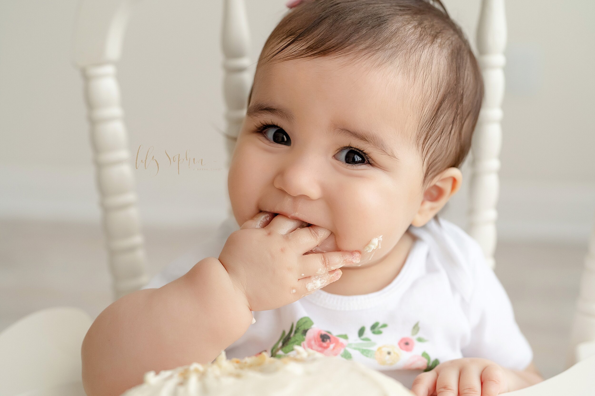  First birthday picture of a little girl licking her icing caked fingers during her cake smash session as she sits in an antique high chair taken in a studio in the Ponce City Market area of Atlanta. 