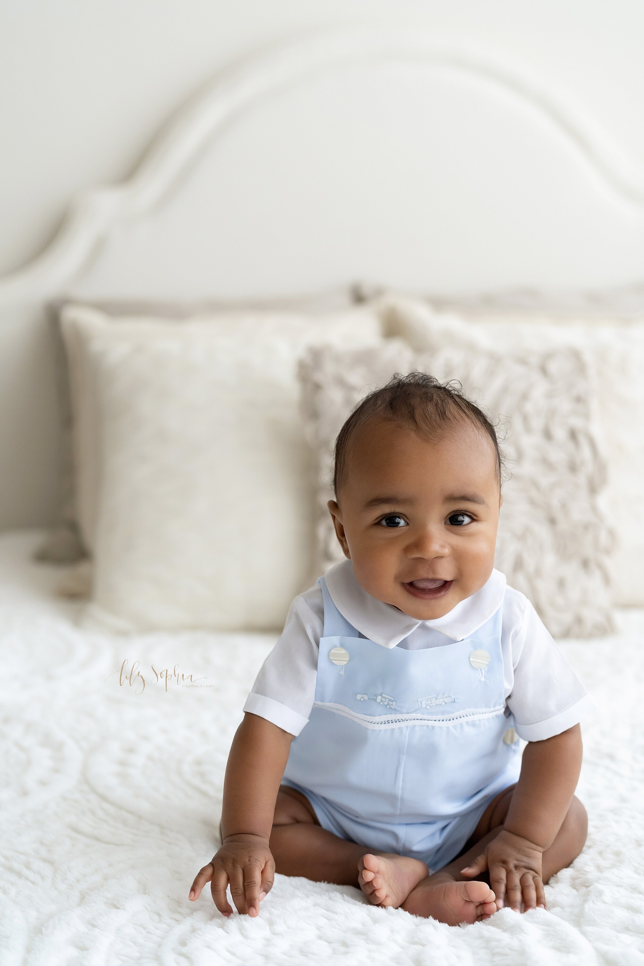  Baby picture of a six month old African-American boy as he sits on top of a bed in an adorable light blue romper and a white peter pan collared shirt taken in a studio near the Poncey Highlands area of Atlanta, Georgia in natural light. 