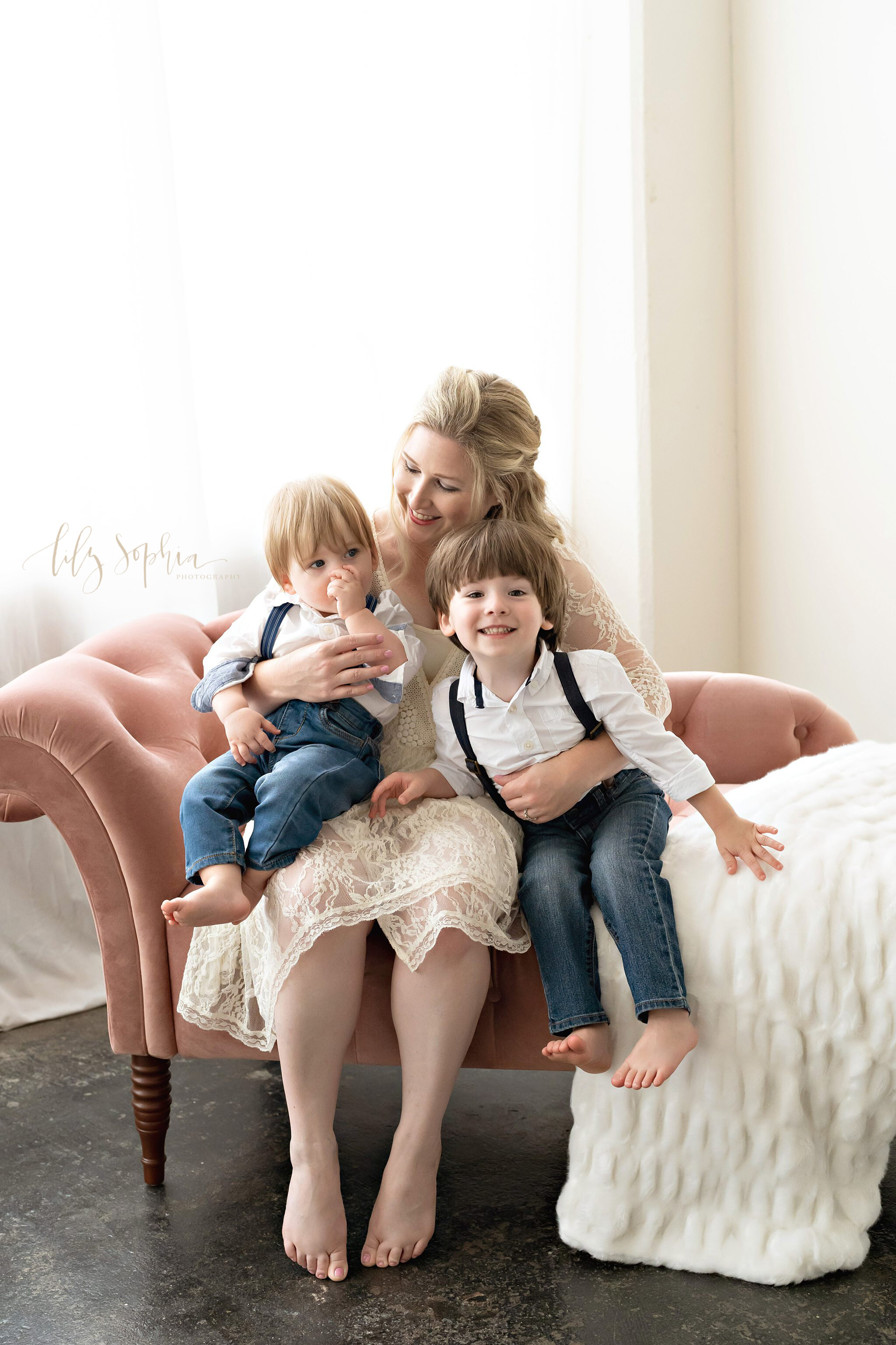  Natural family portrait of a mom holding her young son who is sucking his thumb on her lap while her other son sits next to her on a tufted divan in a natural light studio near the Virginia Highlands area of Atlanta, Georgia. 