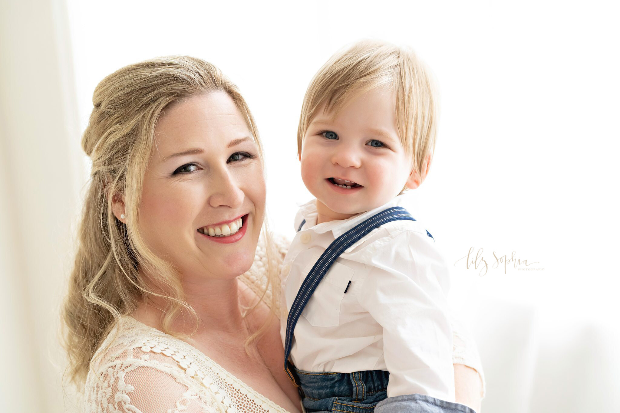  Family photo of a mom holding her young son dressed in blue pants, suspenders, and a long sleeved white shirt as she stands in front of a window in a natural light studio near the Ponce City Market area of Atlanta. 