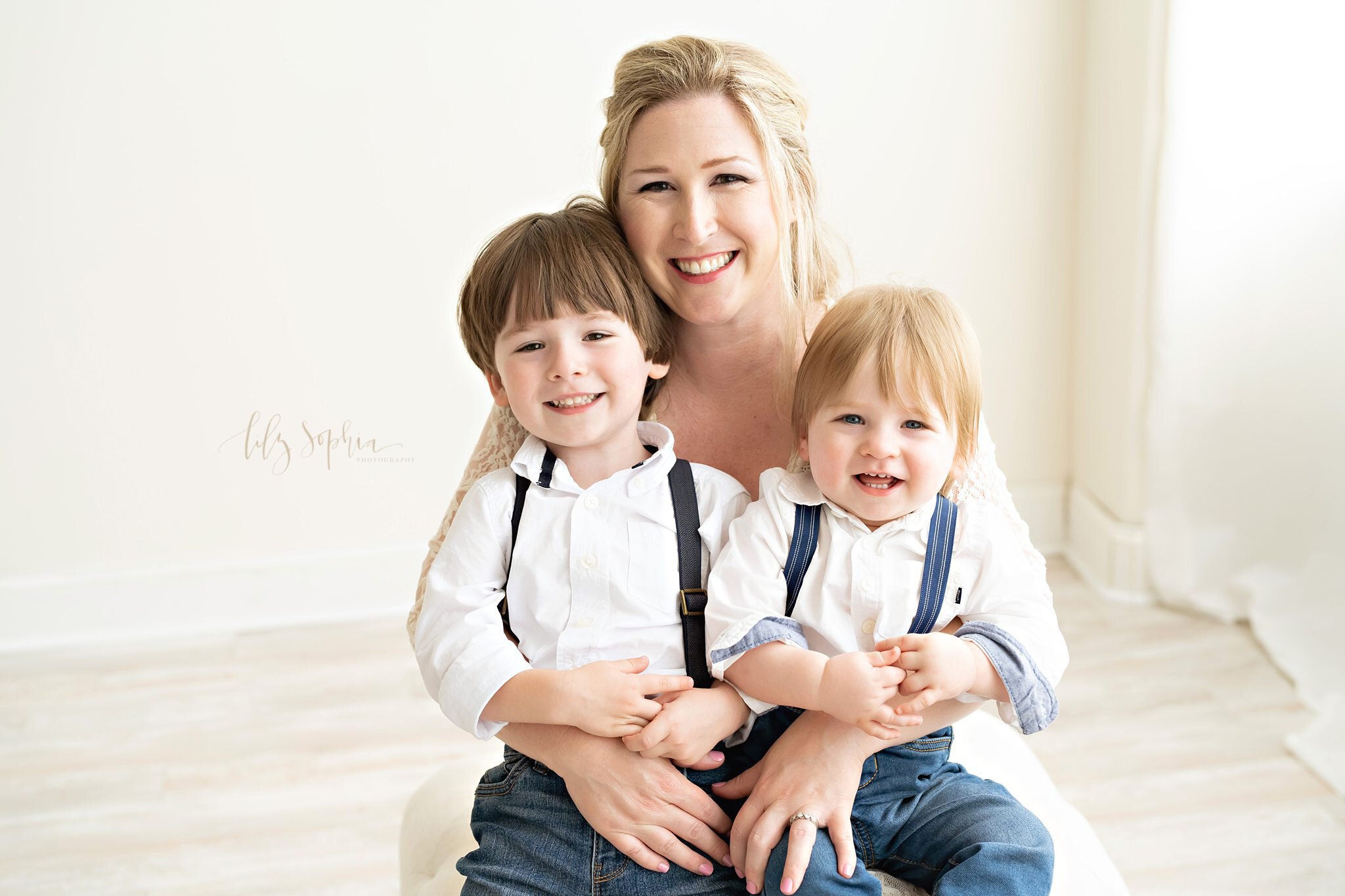  Family portrait of a mom holding her two sons on her lap as she sits on an ottoman in front of a window streaming natural light in a studio near the Midtown area of Atlanta, Georgia. 
