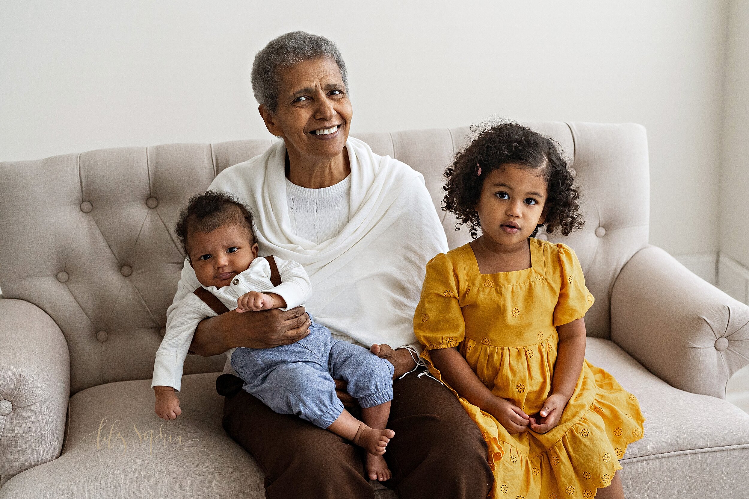  Family photo shoot with an African-American grandmother holding her three month old grandson on her lap while her granddaughter sits next to her on a tufted sofa in a studio near Midtown in Atlanta captured using natural light. 