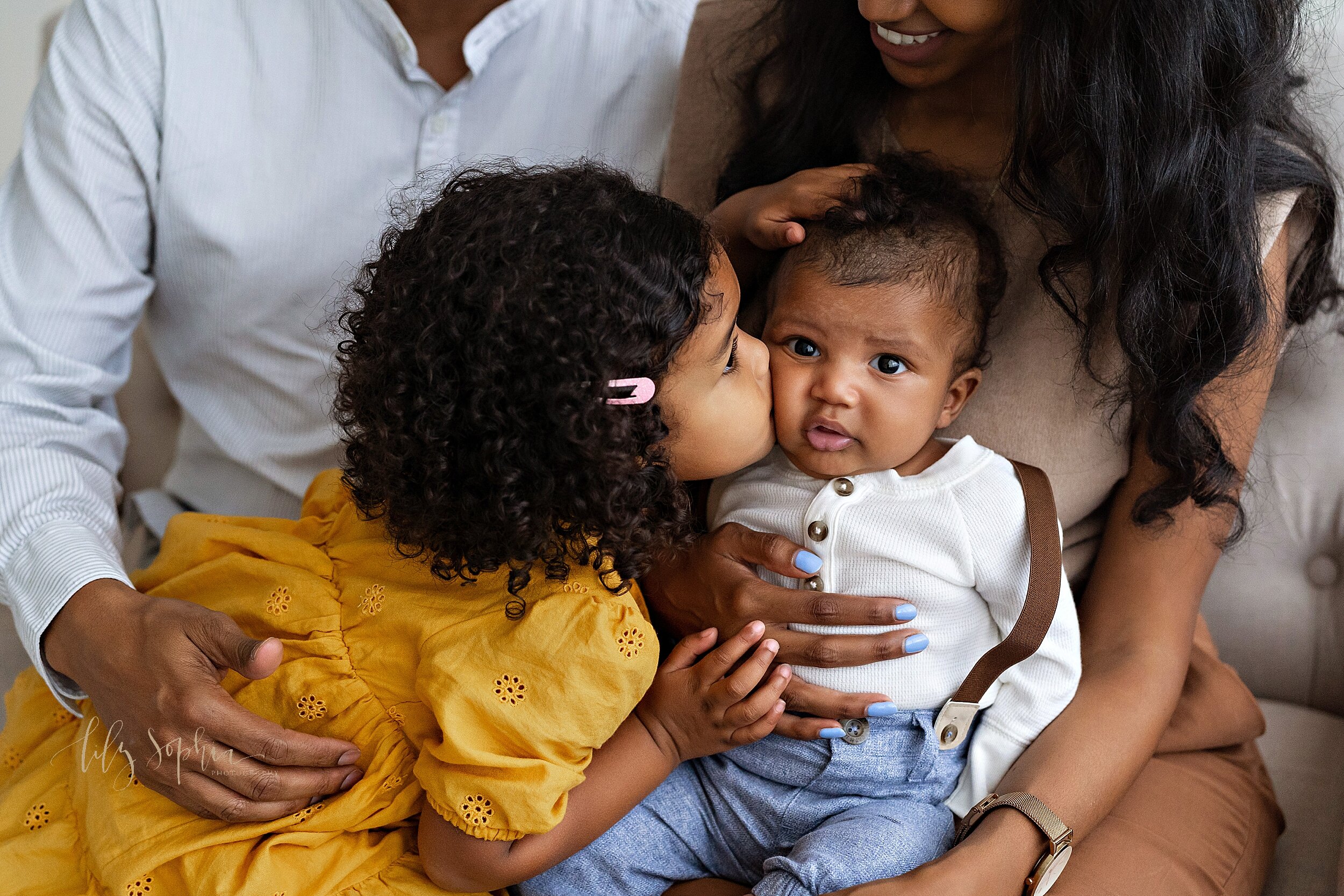  Family photo of an African-American family as dad holds their toddler daughter on his lap and mom holds their three month old baby son on her lap capturing the daughter kissing her baby brother on the cheek taken in a studio near Morningside in Atla