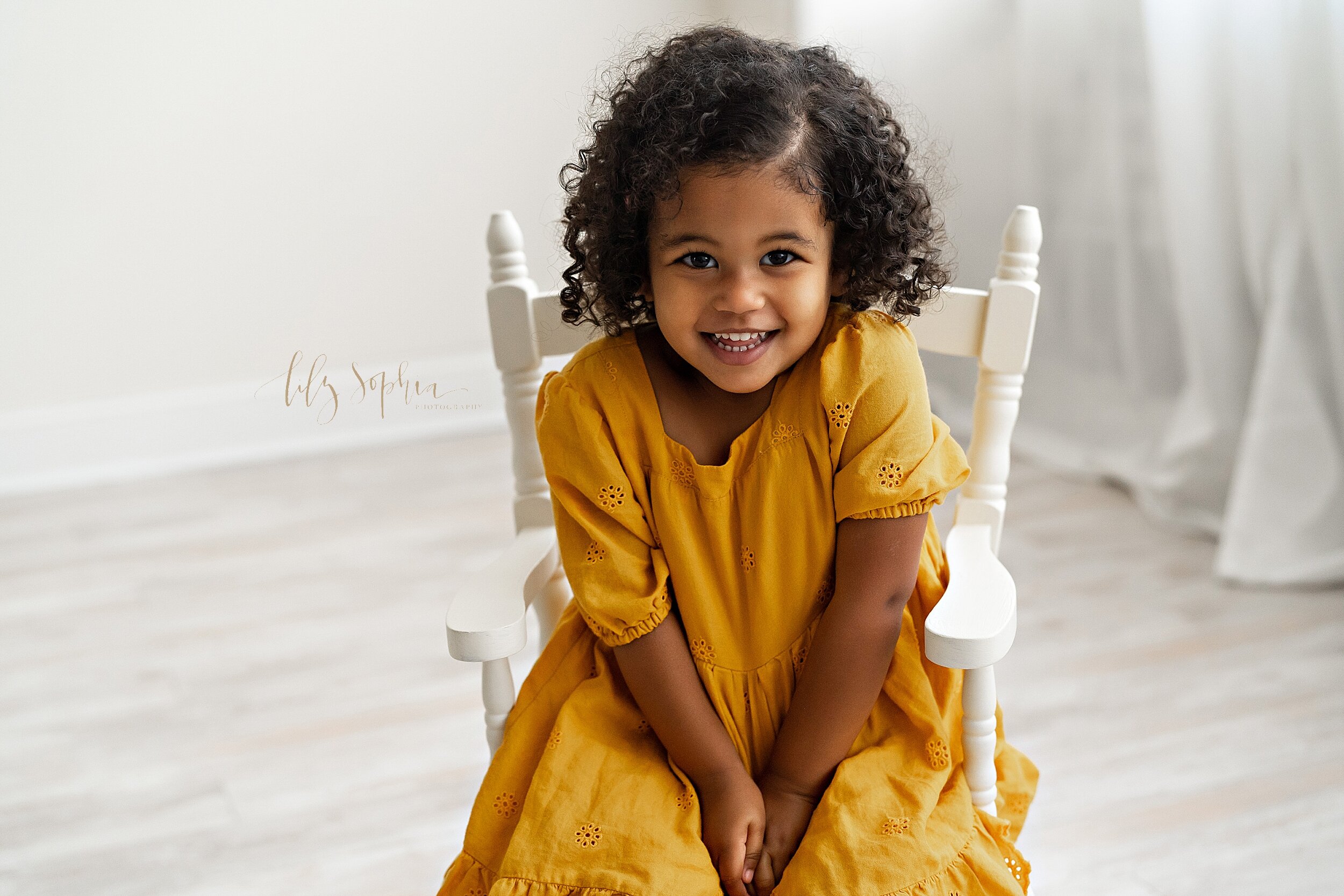 Family photo of an African-American girl as she sits in a white wooden chair in front of a window streaming natural light in a studio near the Candler Park area of Atlanta. 