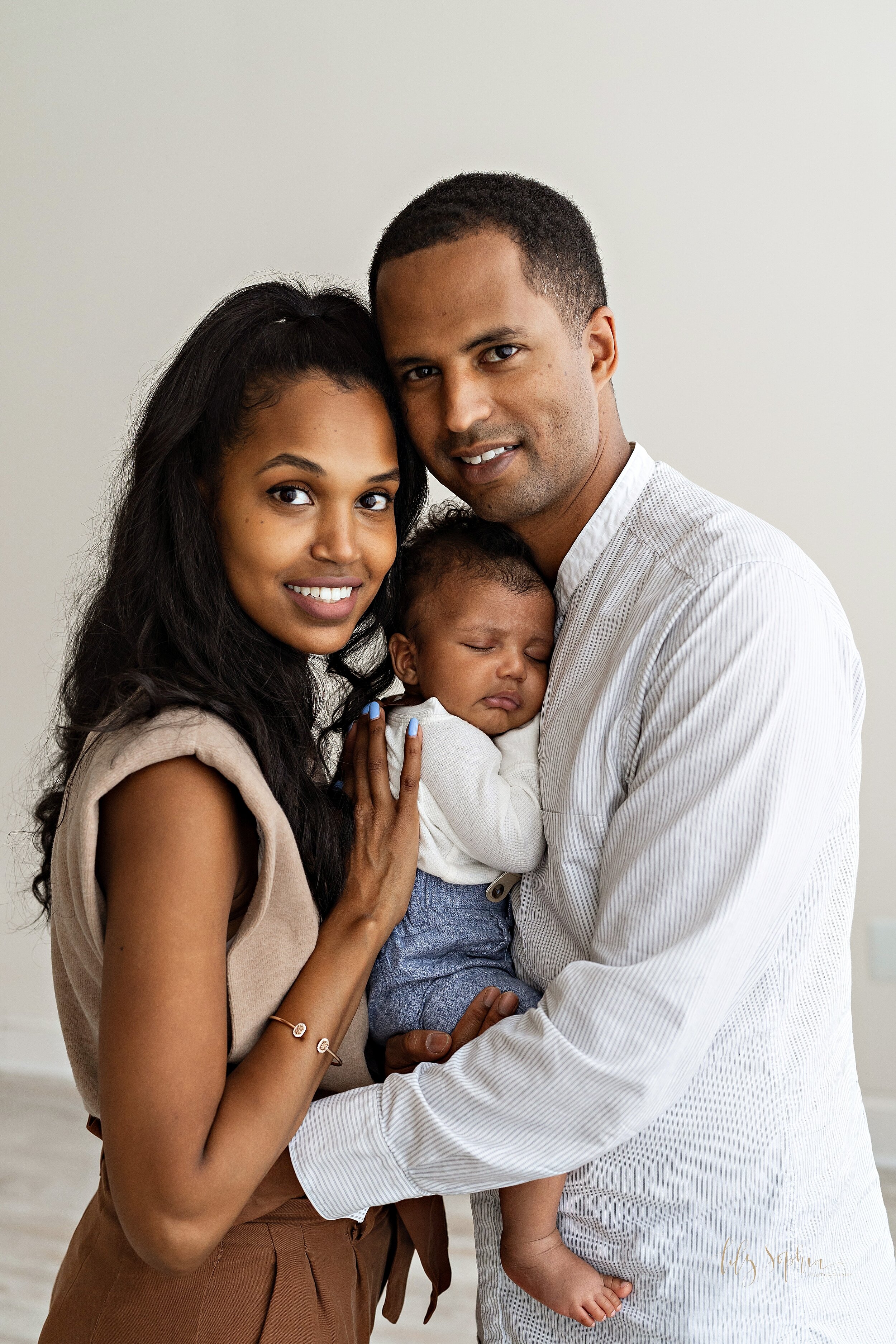  Family portrait of an African-American family as the father holds his sleeping three month old son against his chest and the mother faces the father and places her hands on her son’s back while the two of them stand in a natural light studio near Bu