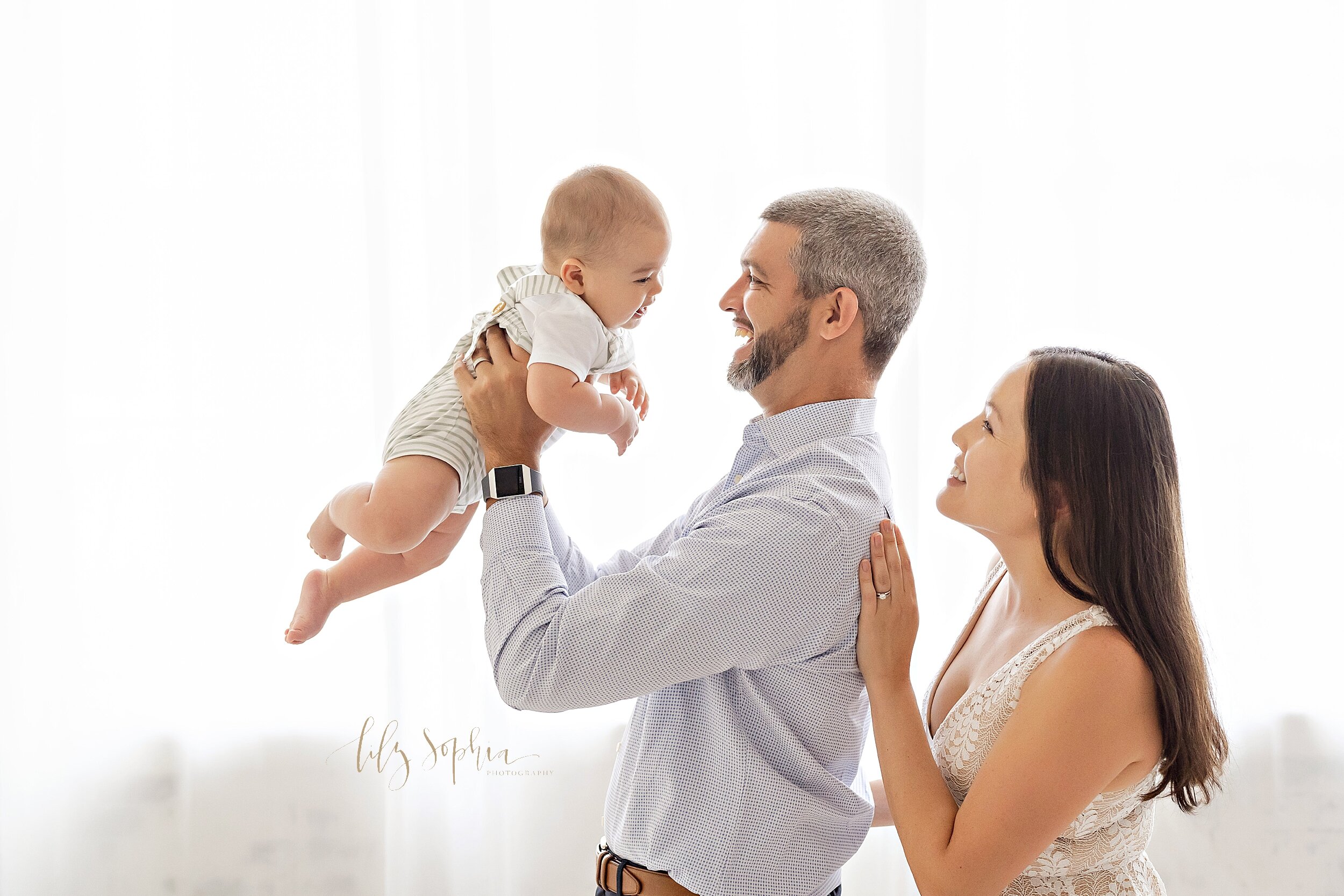  Family photo of a father holding his son in front of him as he stands in front of a window with natural light streaming in and his wife stands behind her husband with her hands against his back as they smile at their son taken in a studio near the B