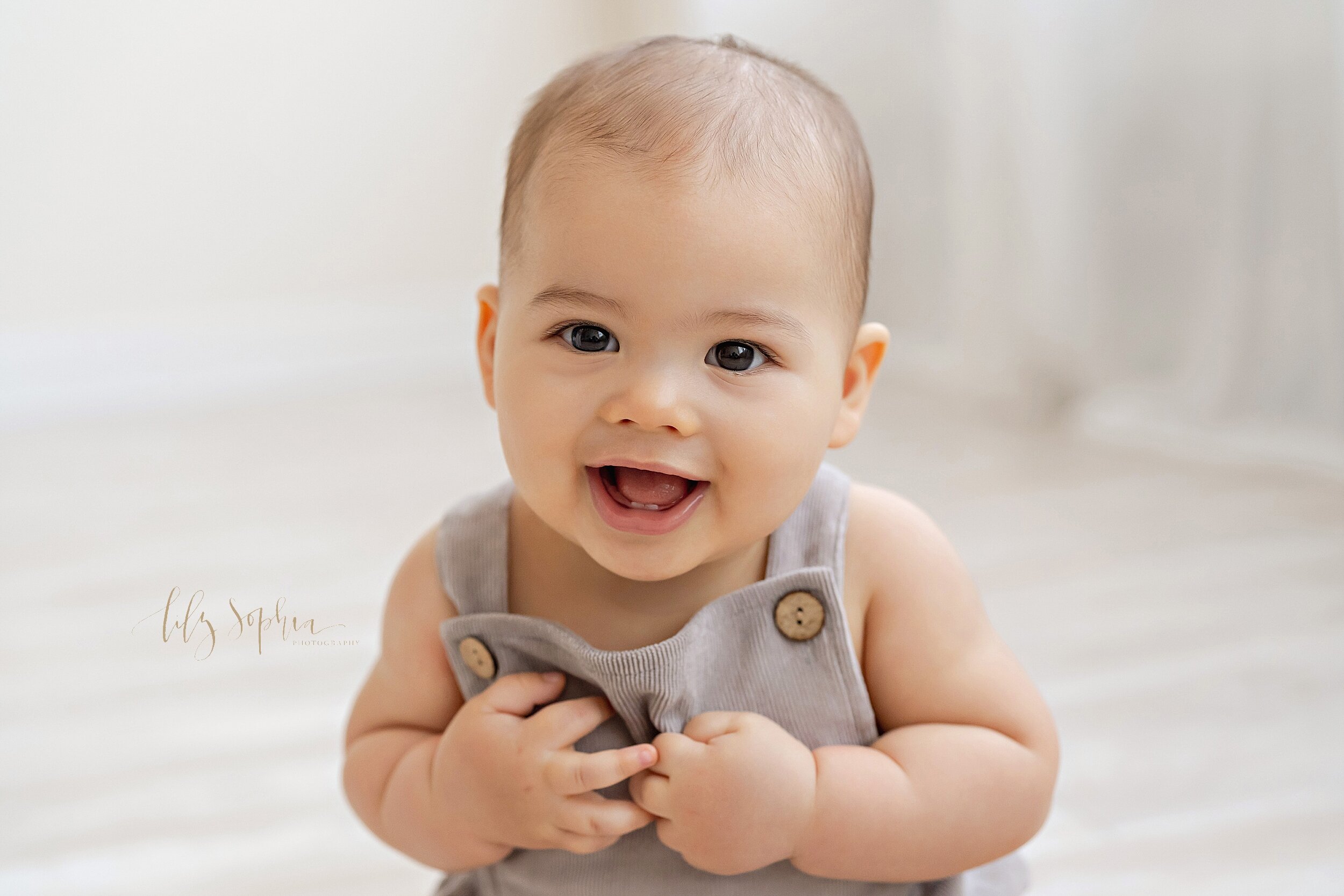  Baby photo of a six month old little boy as he sits in a studio, points to himself, and smiles to show his two little teeth in his mouth taken in a natural light studio near the Decatur area of Atlanta. 