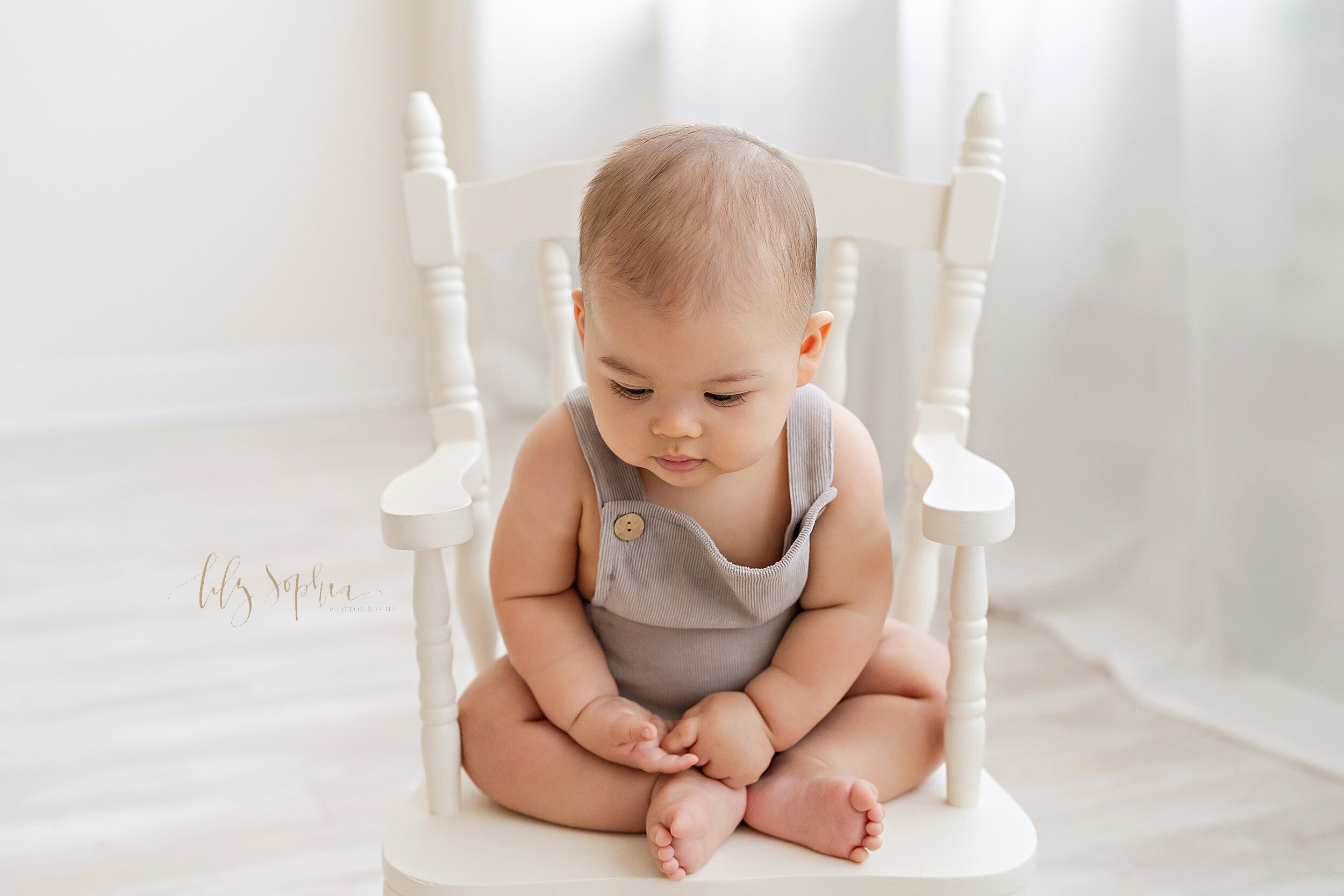 Baby photo of a six month old little boy as he sits in a white rocking chair and looks down to the floor taken near the Kirkwood area of Atlanta in a natural light studio. 