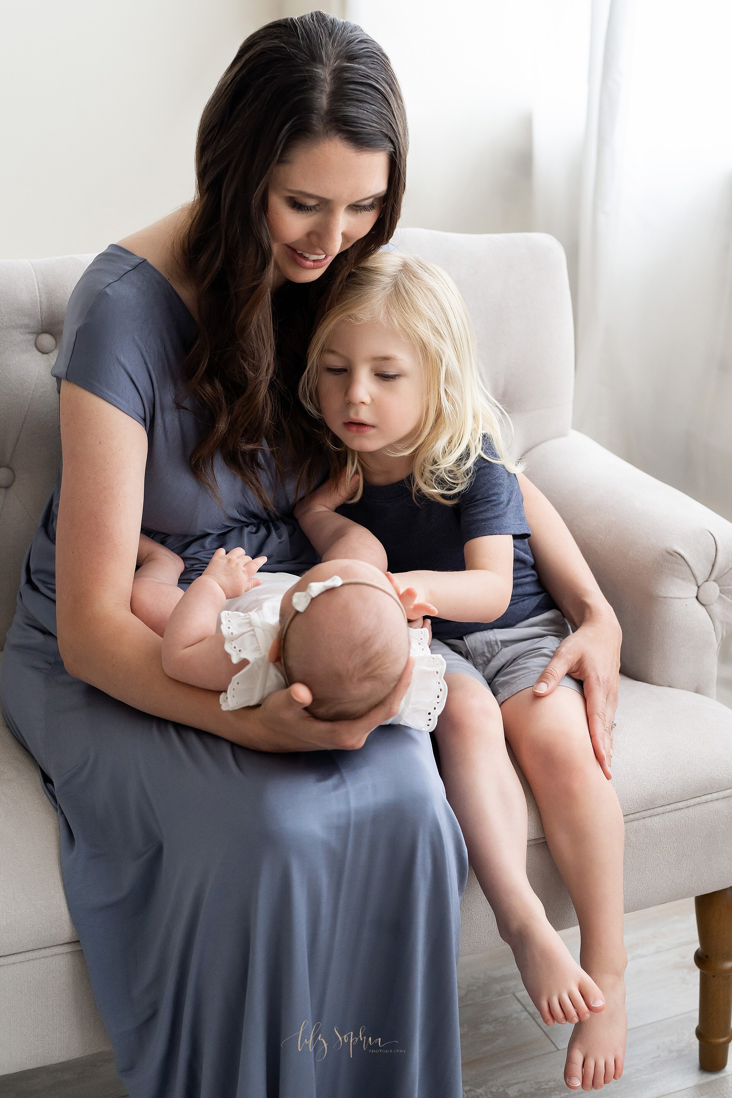  Family photo of a mom dressed in a blue full-length gown holding her three month old daughter on her lap while her son sits next to her on a tufted sofa in a natural light studio near Decatur in Atlanta. 