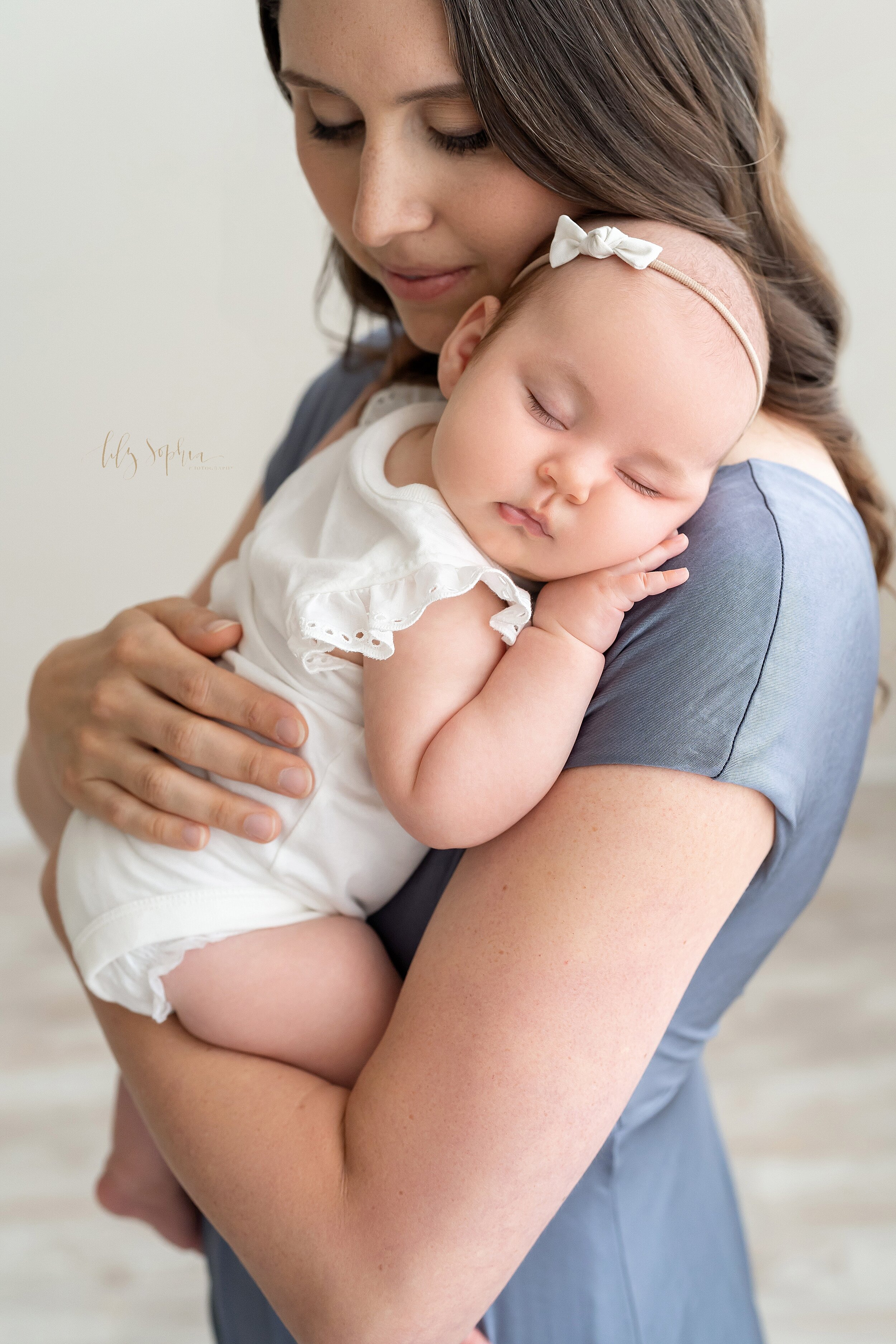  Family photo of a mom holding her sleeping three month old daughter on her shoulder as she stands in a studio near Midtown in natural light. 