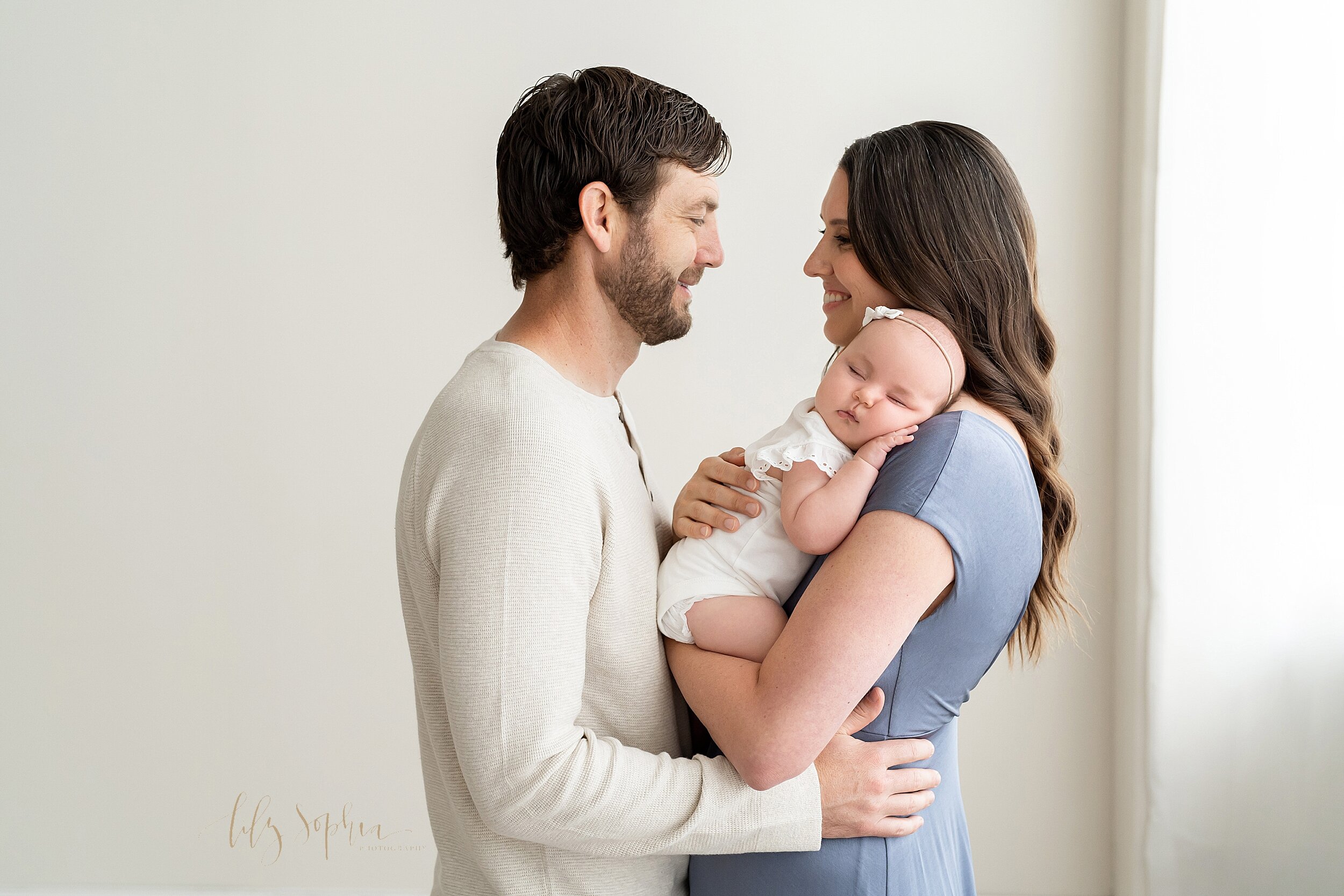  Family picture of a father facing his wife with his hands on her waist as she holds their sleeping three month old daughter on her shoulder and the two of them look lovingly at one another taken in a natural light studio near Morningside in Atlanta,