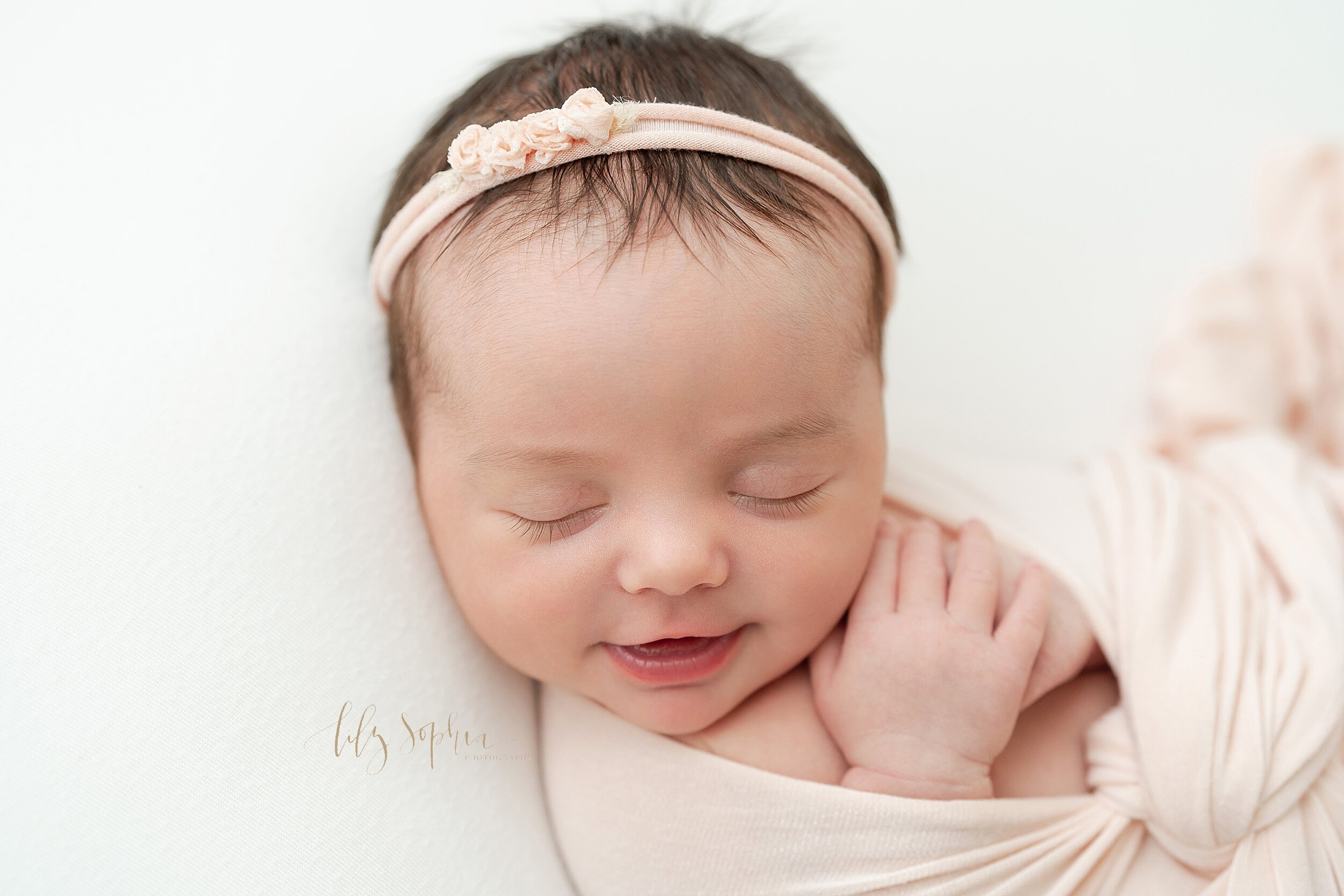  Newborn photo of a sleeping and smiling baby girl as she sleeps with a stretchy headband in her wispy hair and wrapped in a stretchy swaddle taken in a studio near the Old Fourth Ward area of Atlanta in natural light. 