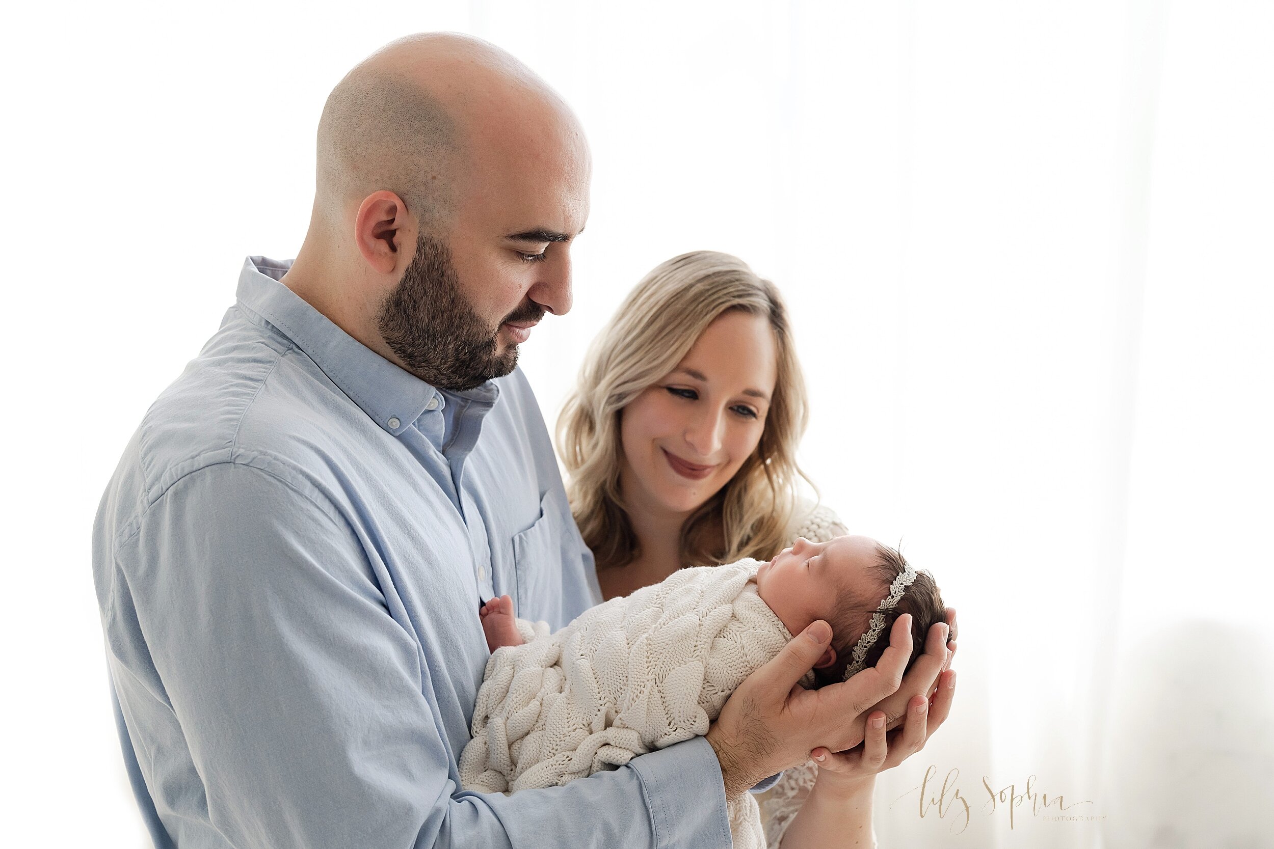  Family photo of a father standing in front of window streaming natural light and holding his newborn daughter in his hands as his wife smiles at their daughter as she stands to her husband’s left taken in Ponce City Marker in the Lily Sophia Photogr