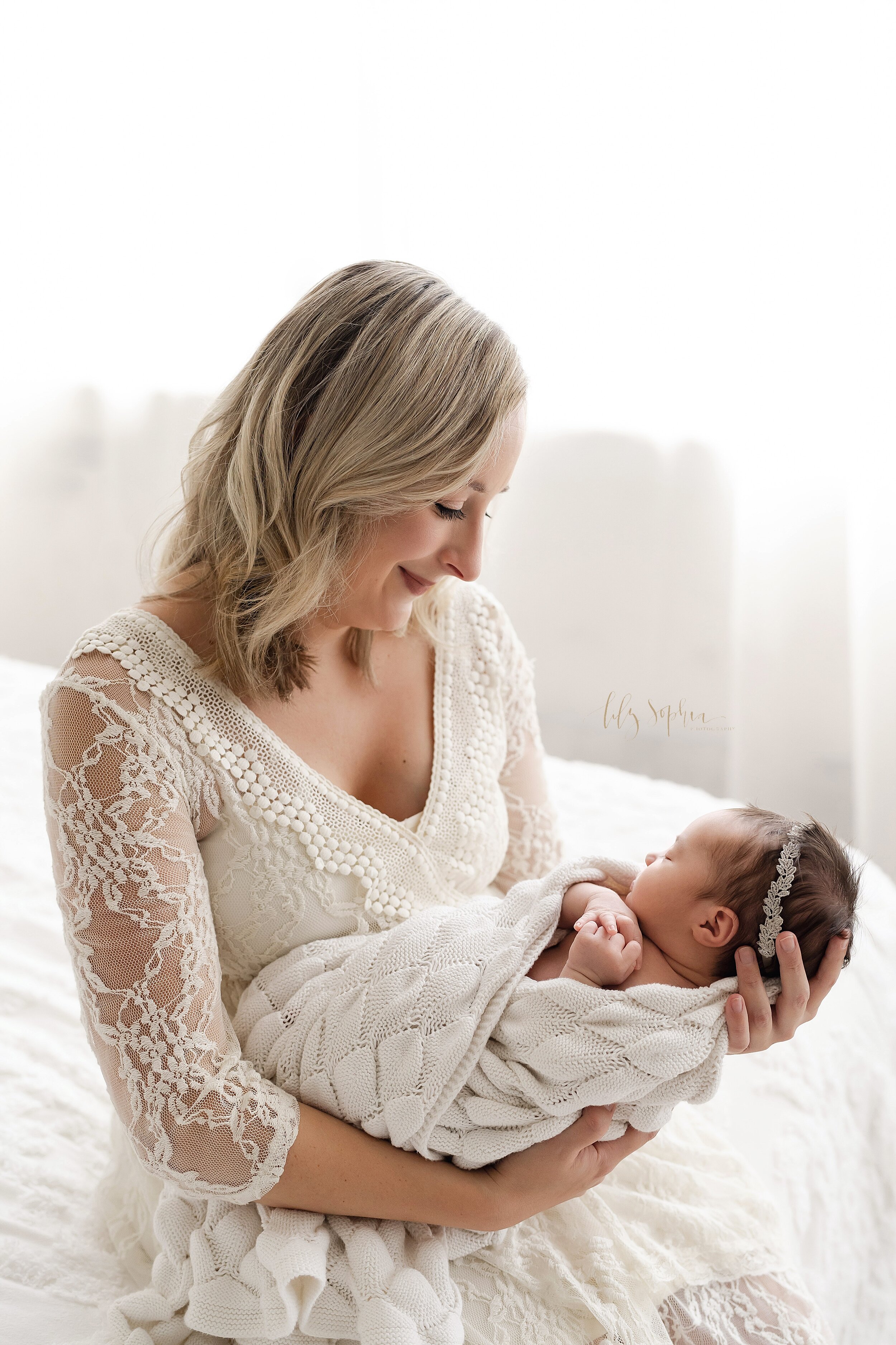  Family photo of a mother holding her sleeping newborn baby girl in her arms as she sits on a bed in front of a window streaming natural light in a studio near Midtown in Atlanta, Georgia. 