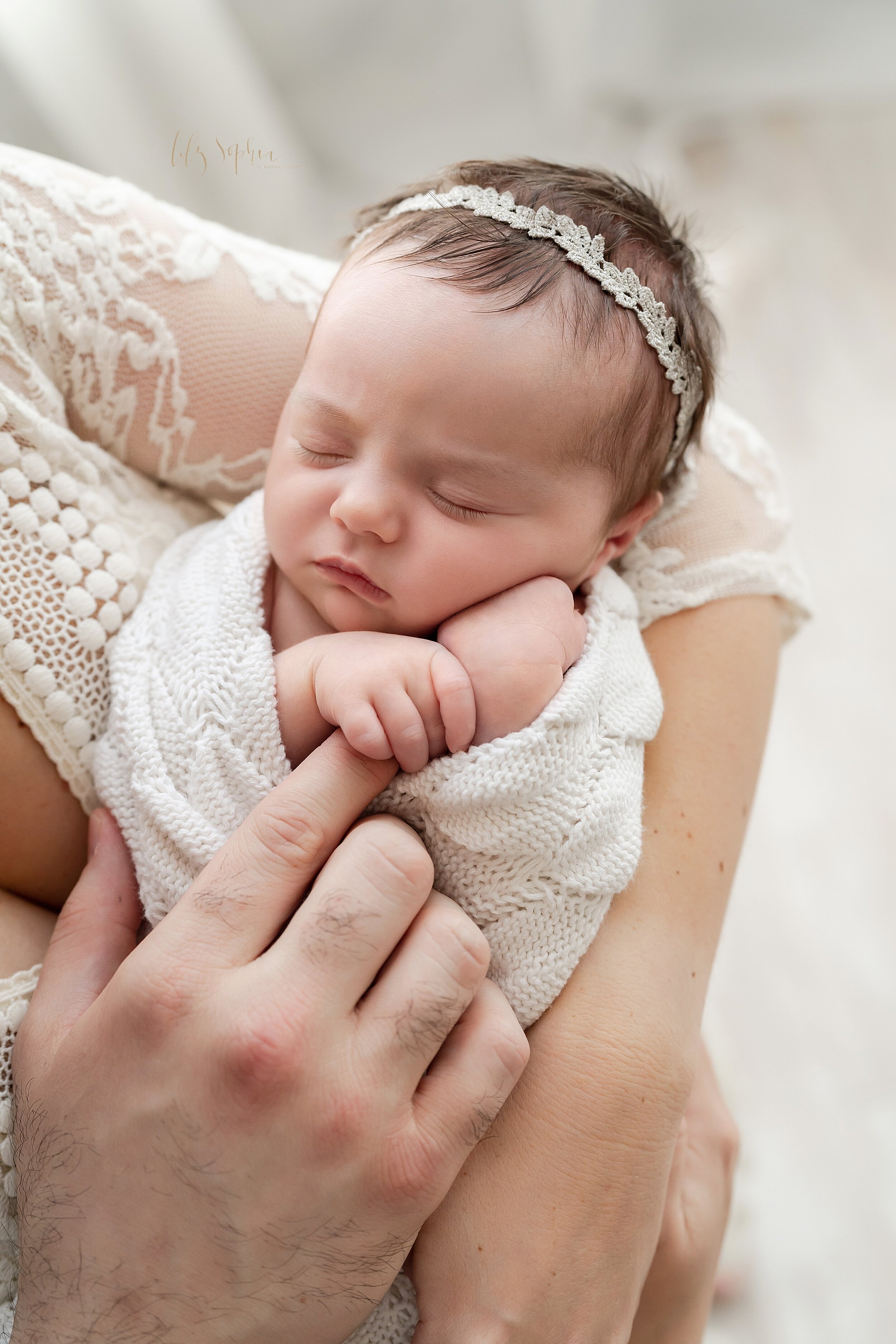 Close-up picture of a peacefully sleeping infant girl being held by her mother as the baby girl holds the pointer finger of her father taken in a studio near Alpharetta in Atlanta in natural light. 