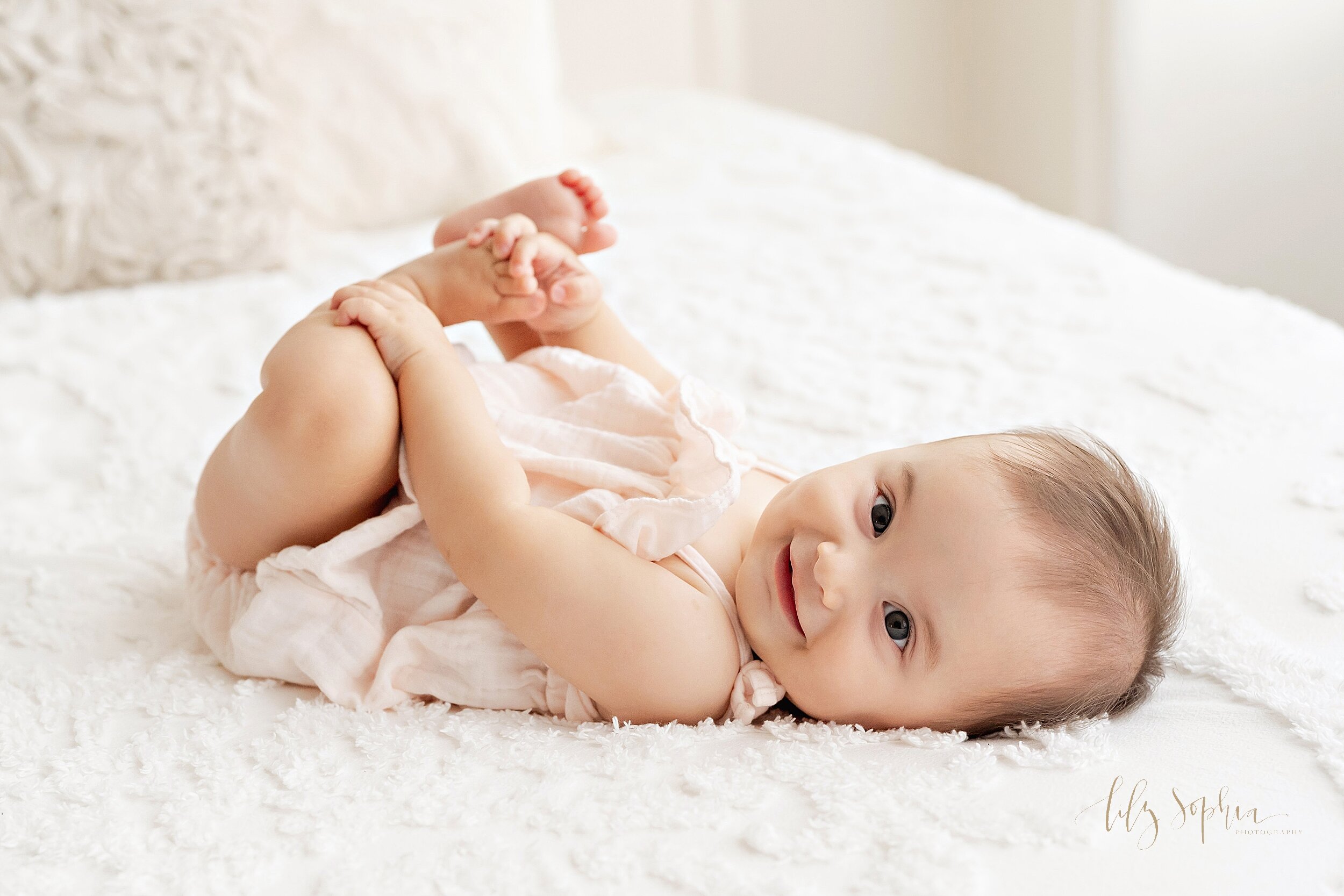  Baby photo of a six month old little girl dressed in a pink romper as she plays with her toes while laying on her back on a bed taken in a studio near Old Fourth Ward in Atlanta, Georgia in natural light. 