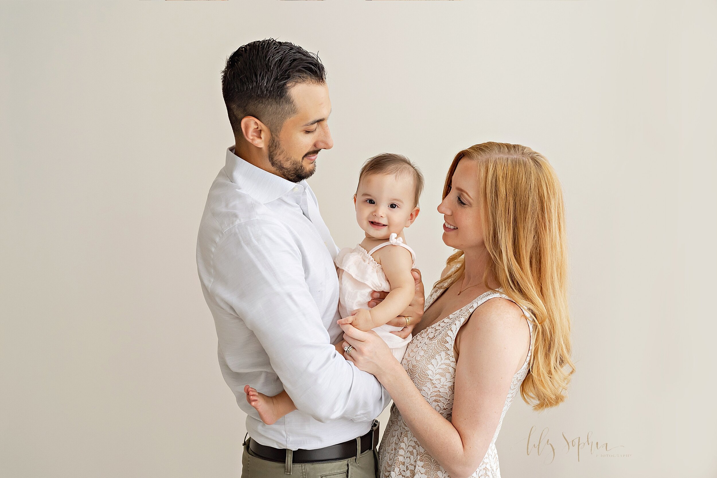  Family photo of a dad holding his six month old daughter at his waist while mom stands facing dad and the two of them admire their smiling daughter taken in a natural light studio near Morningside in Atlanta. 