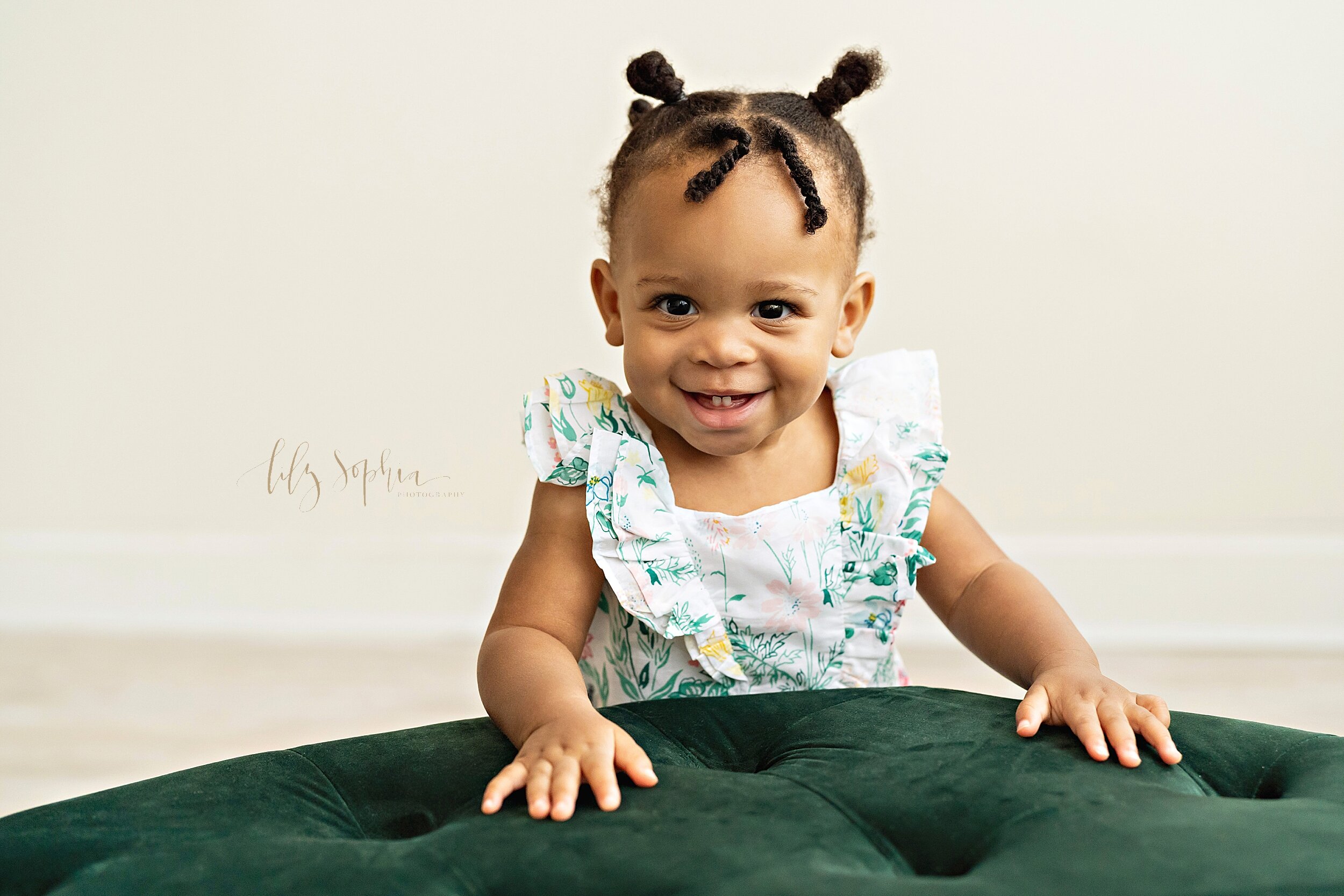 Baby photo of an African-American one-year old girl as she stands with her hands on the top of a tufted ottoman in a studio near Poncey Highlands in Atlanta in natural light. 