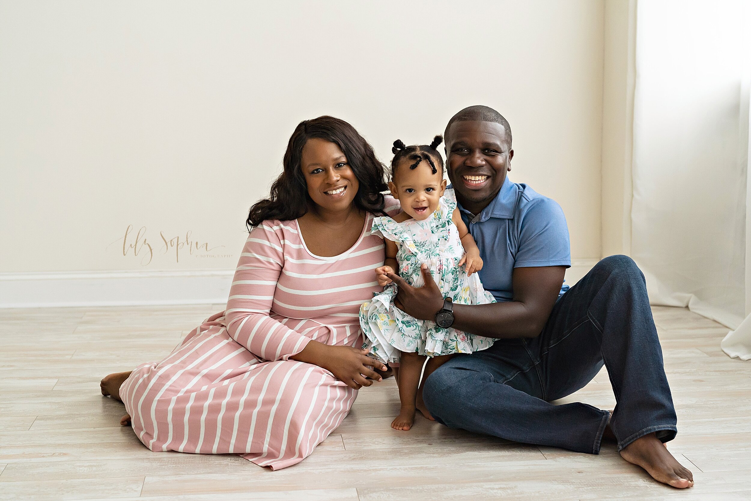  Family photo of a happy African-American mom and dad sitting on the floor of a natural light studio with their one-year old daughter standing between them as dad steadies her taken near Midtown in Atlanta, Georgia. 