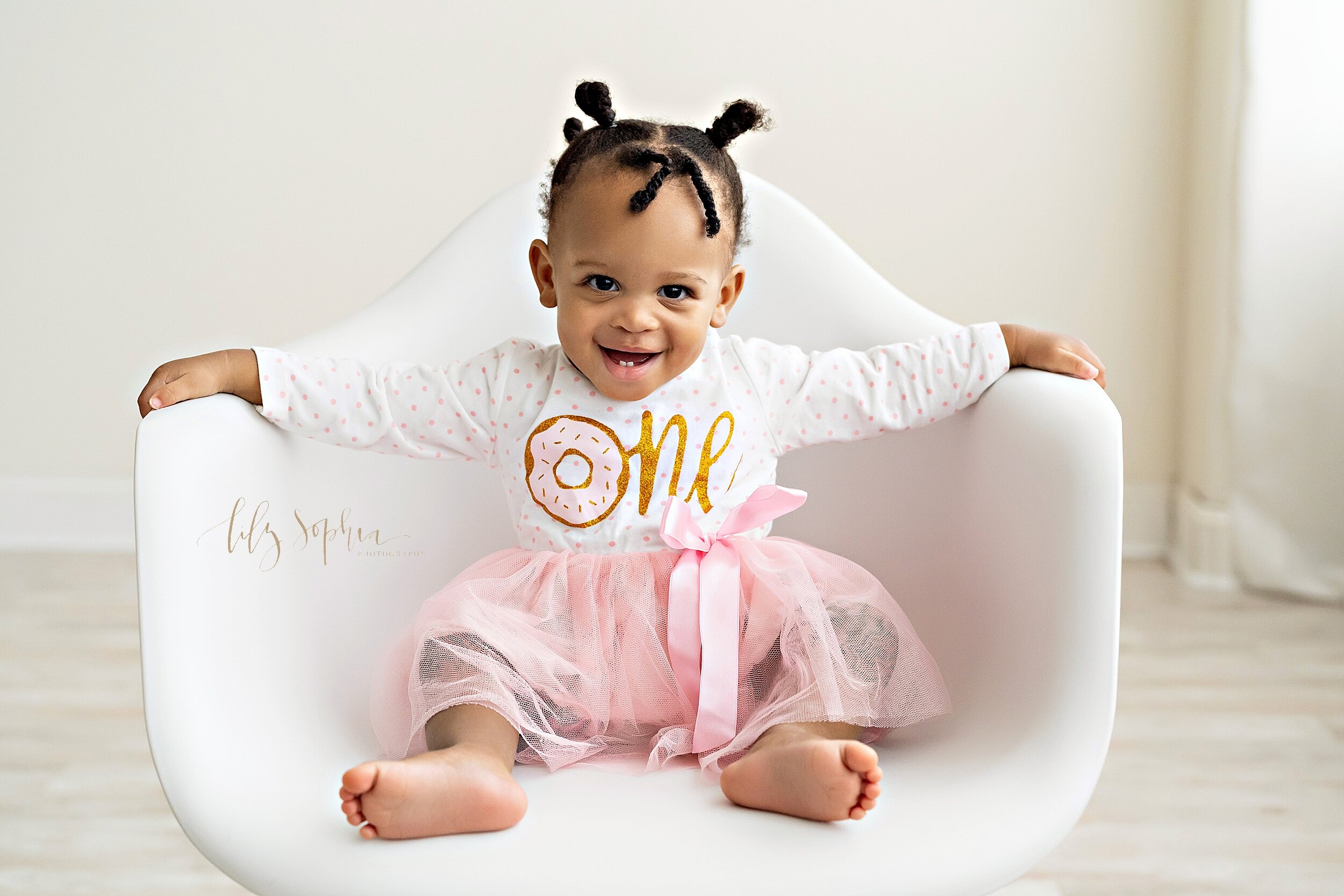  First birthday photo of an African-American girl dressed in a shirt with the word one on it and a pink tutu skirt as she sits in a white molded chair smiling to show her two tiny teeth on the bottom taken in natural light in a studio near Old Fourth