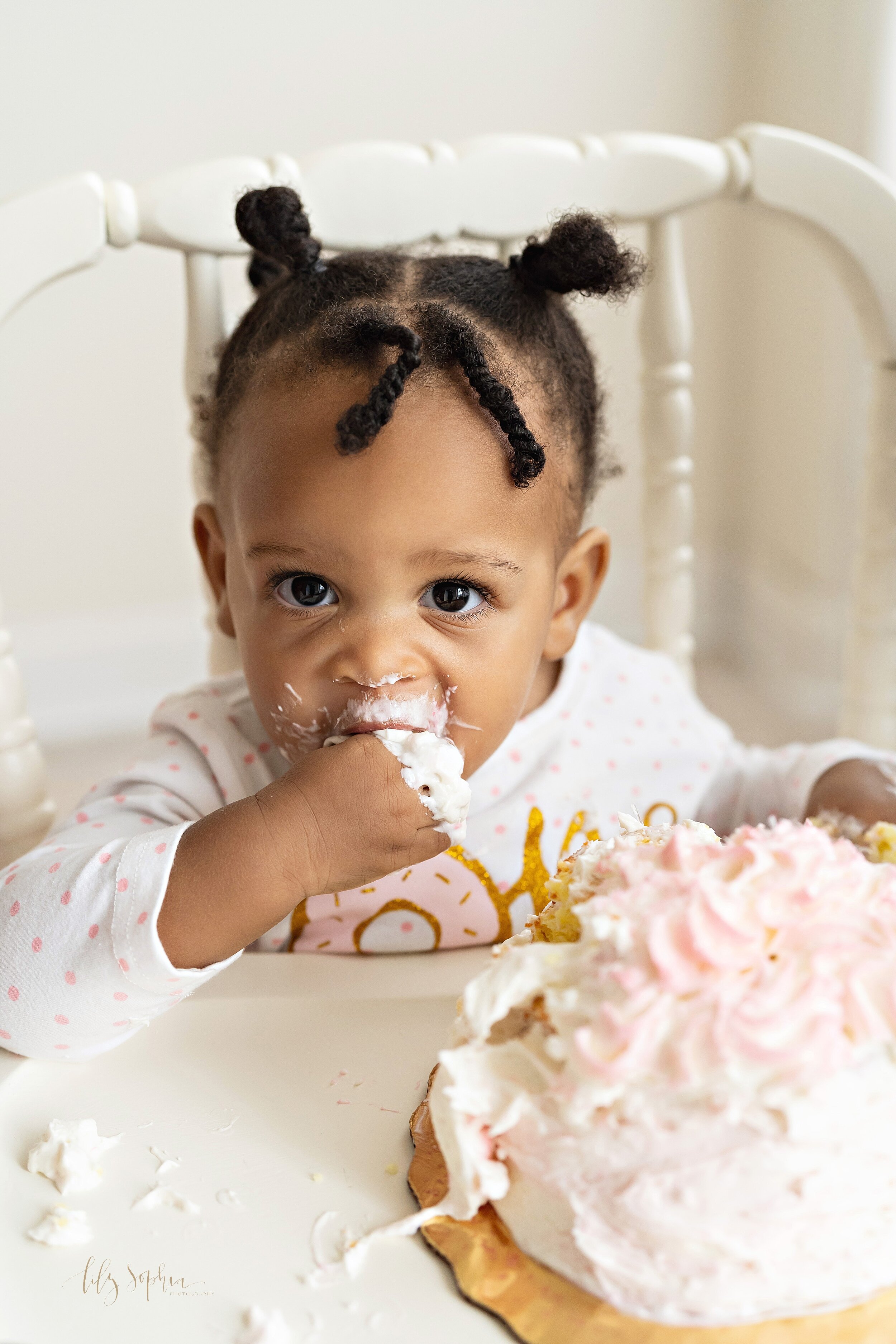  Close-up baby photo of an African-American girl as she stuffs cake and icing into her mouth from her first birthday smash cake taken near Virginia Highlands in Atlanta in a natural light studio. 