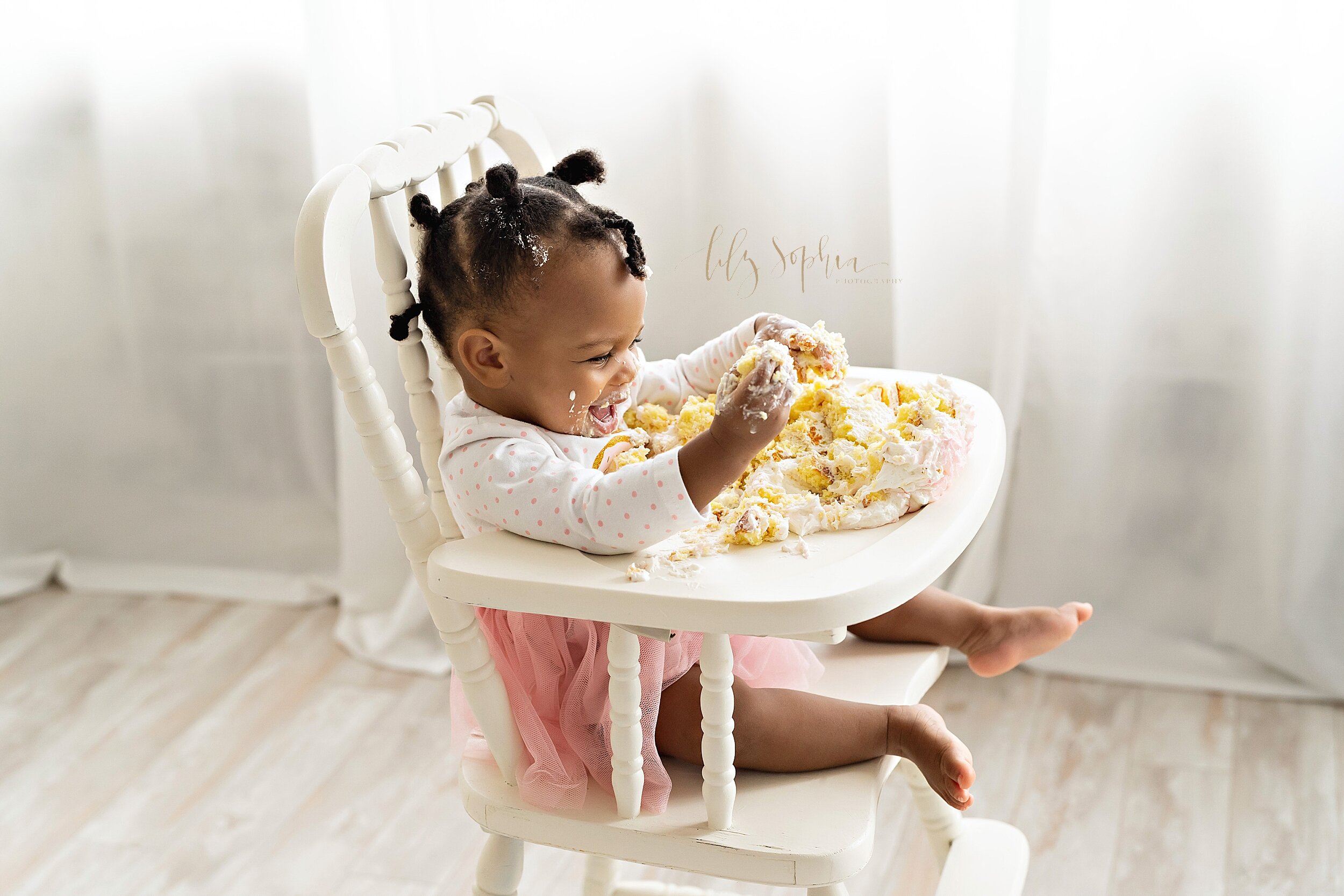  Baby photo of an African-American one year old girl as she smashes her first birthday cake while sitting in an antique highchair in front of a window streaming natural light in a studio near Ponce City Market in Atlanta, Georgia. 