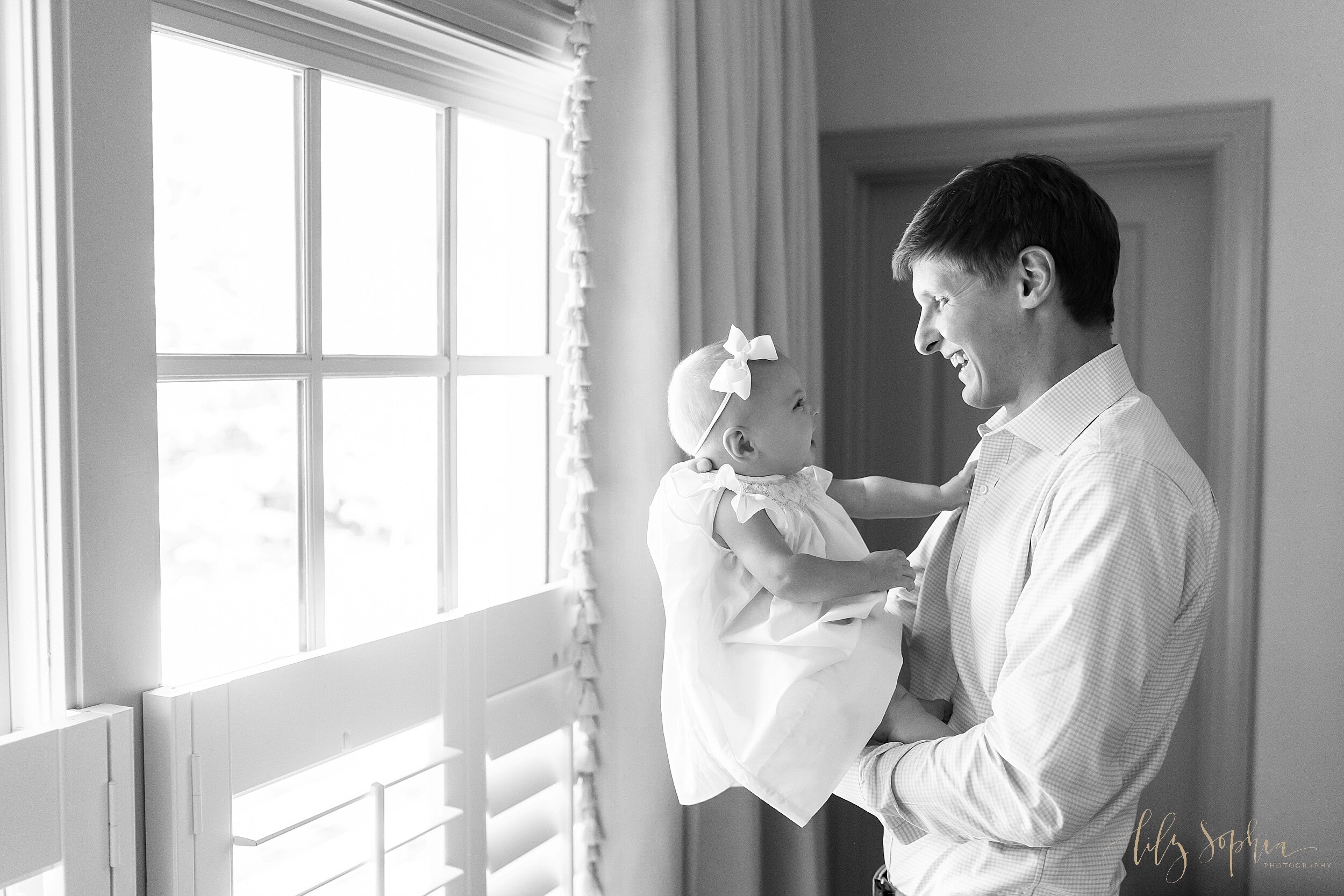  Family lifestyle photo of a father holding his six month old daughter in front of a window in their home near Atlanta as the two of them laugh together. 