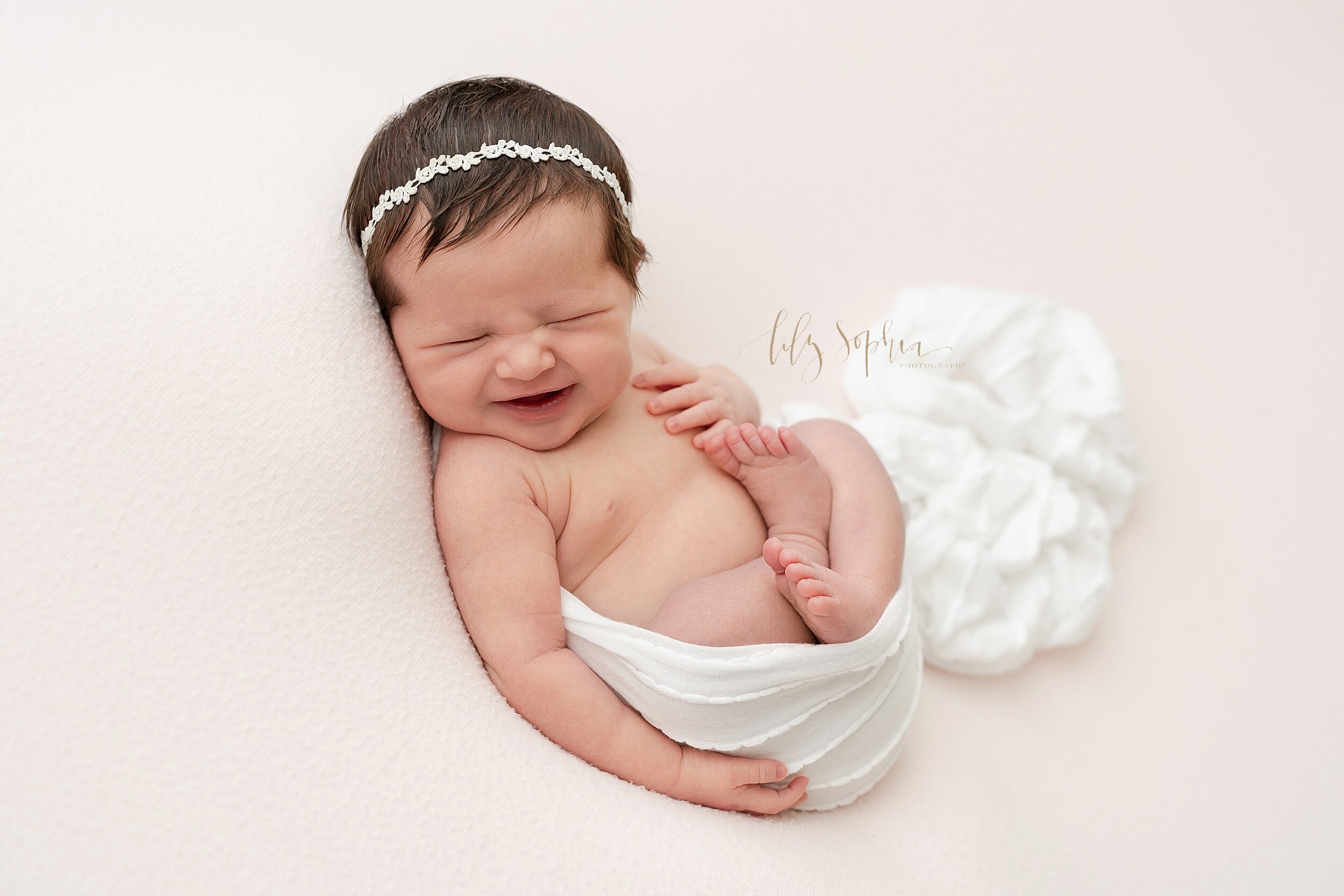  Precious newborn photo of a laughing infant girl as she lies on her back in a stretchy swaddle wearing a lacey laurel leaf headband taken in a studio in Ponce City Market in Atlanta, Georgia in natural light. 