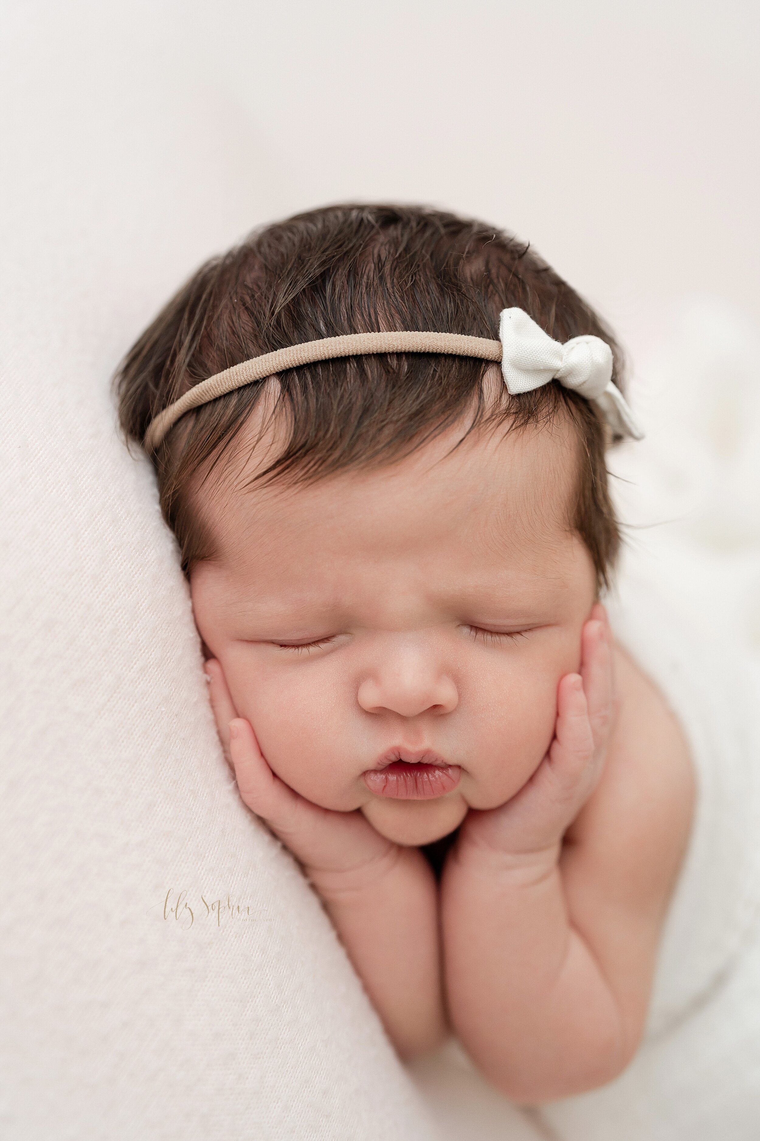  Close-up newborn photo of an infant girl wearing a bow headband in her hair as she holds her cheeks while sleeping on her side in a studio near Candler Park in Atlanta taken in natural light. 