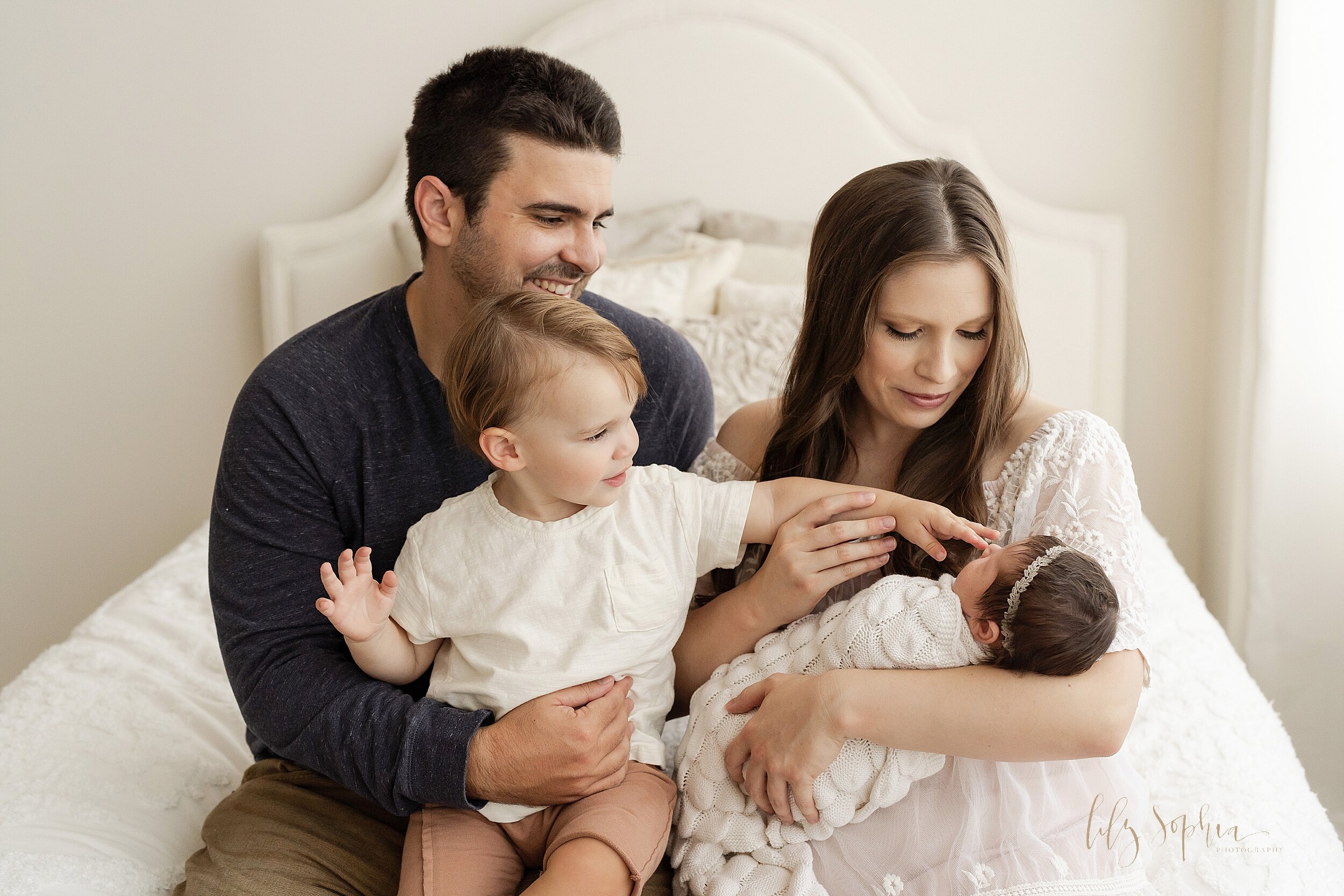  Family newborn photo of a mom holding her newborn daughter in her arms with dad sitting with the couple’s toddler son on his lap as the family sits on the edge of a bed and the toddler son touches his sister’s nose taken in a natural light studio ne