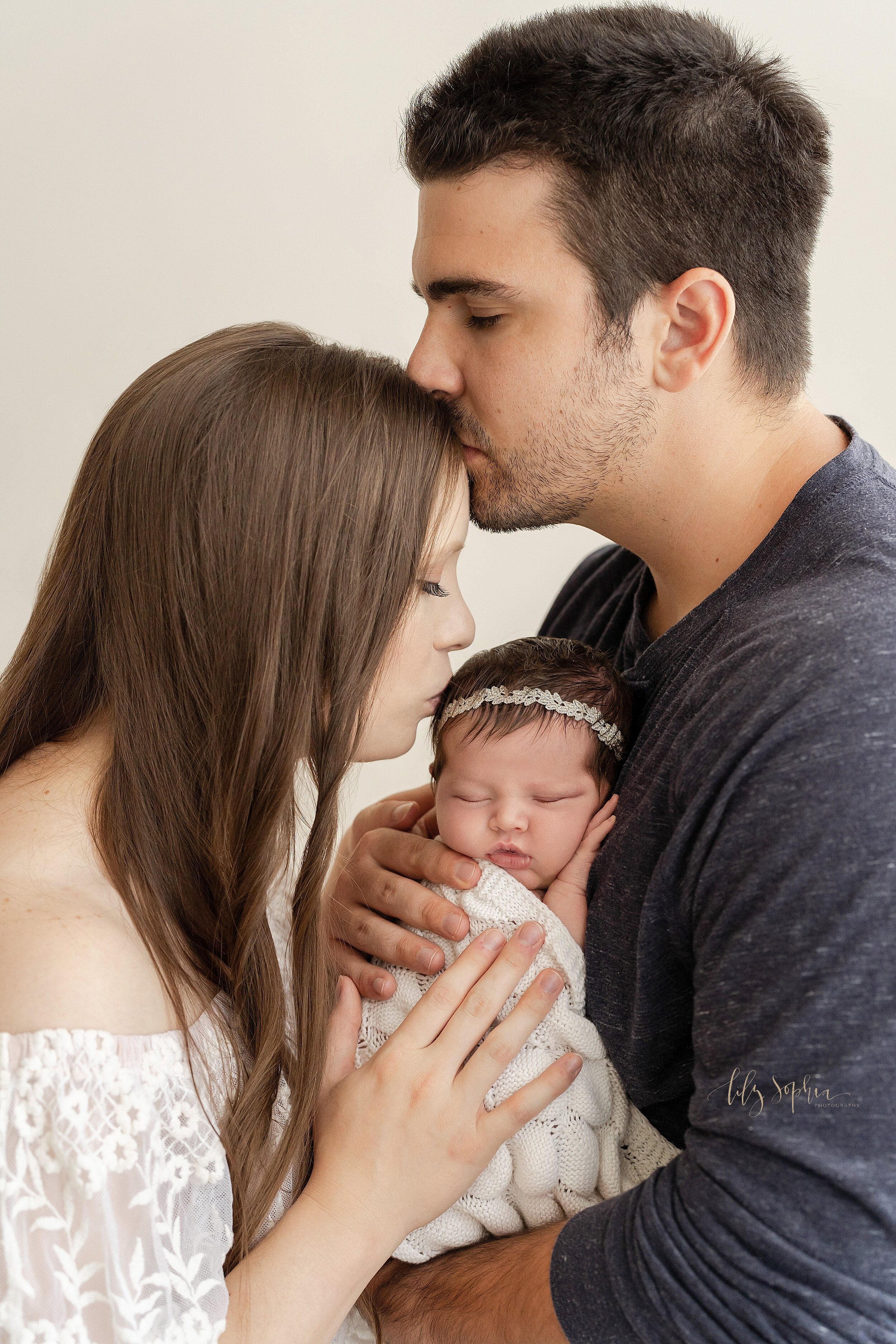  Family newborn photo of a father holding his sleeping newborn daughter against his chest as the mother faces her husband and kisses the side of her daughter’s head while dad kisses his wife’s forehead demonstrating the love in this family taken in a