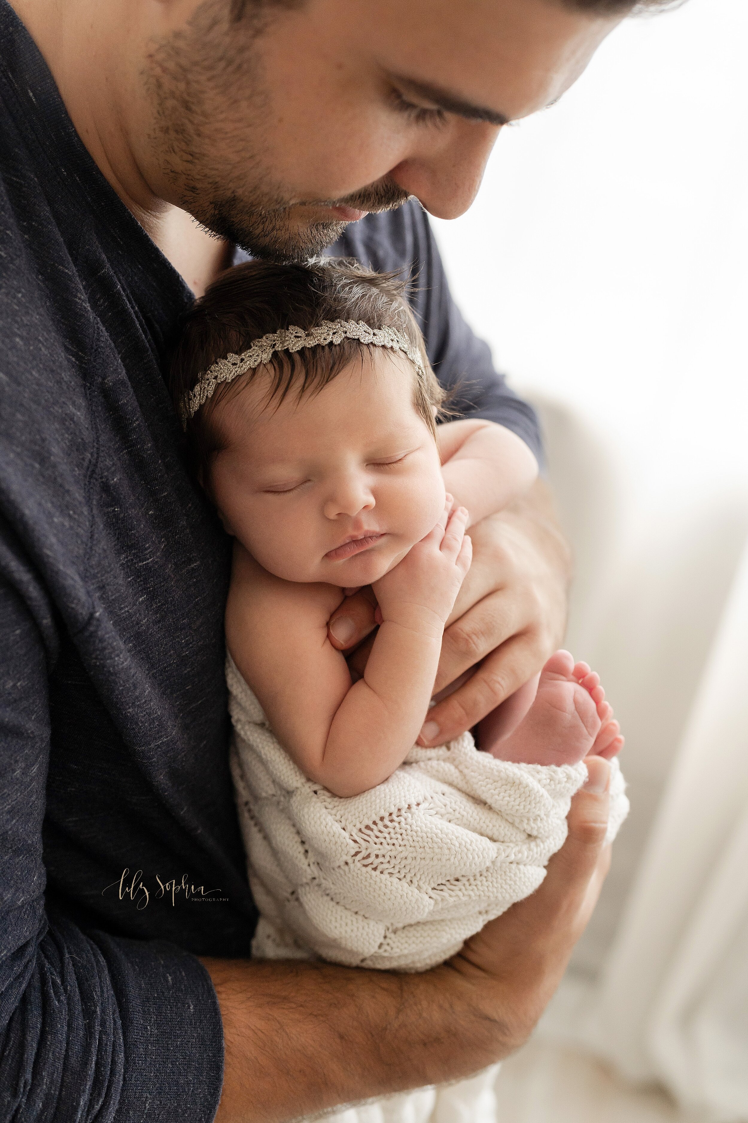  Newborn photo of an infant girl being held in front of her father’s chest with her back to her father as she peacefully sleeps taken in natural light in a studio near Midtown in Atlanta. 