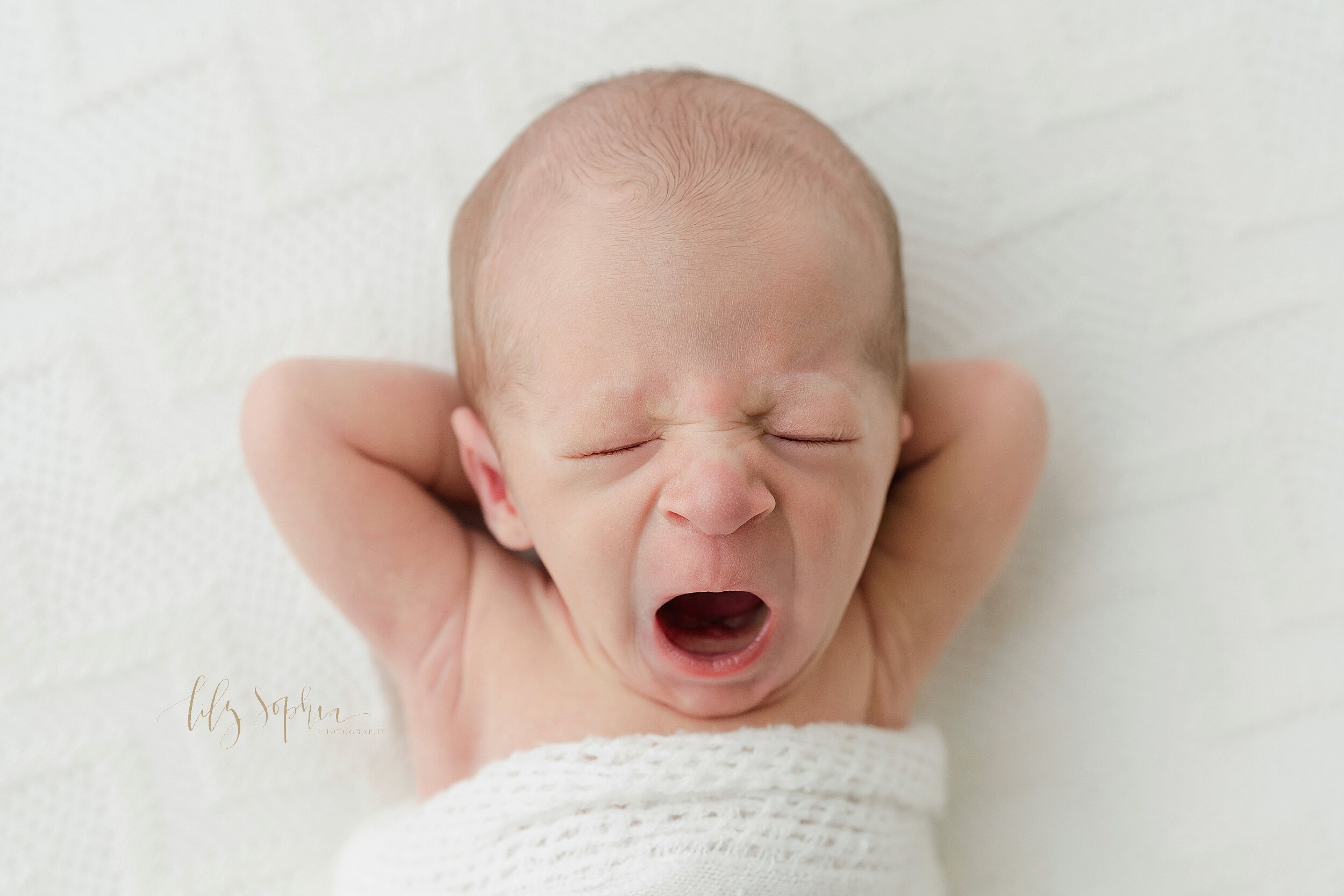  Newborn photo of an infant boy as he lies on his back with his hands behind his head and yawns taken in natural light in a studio near Alpharetta in Atlanta, Georgia. 